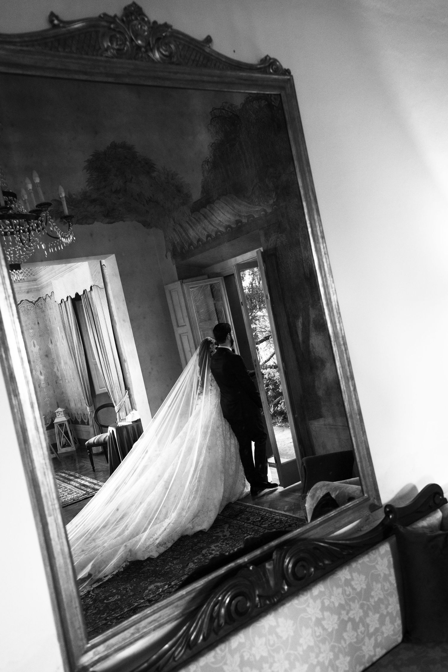 Black and white photo of a bride in a long veil and groom reflected in an ornate antique mirror inside an elegant room.