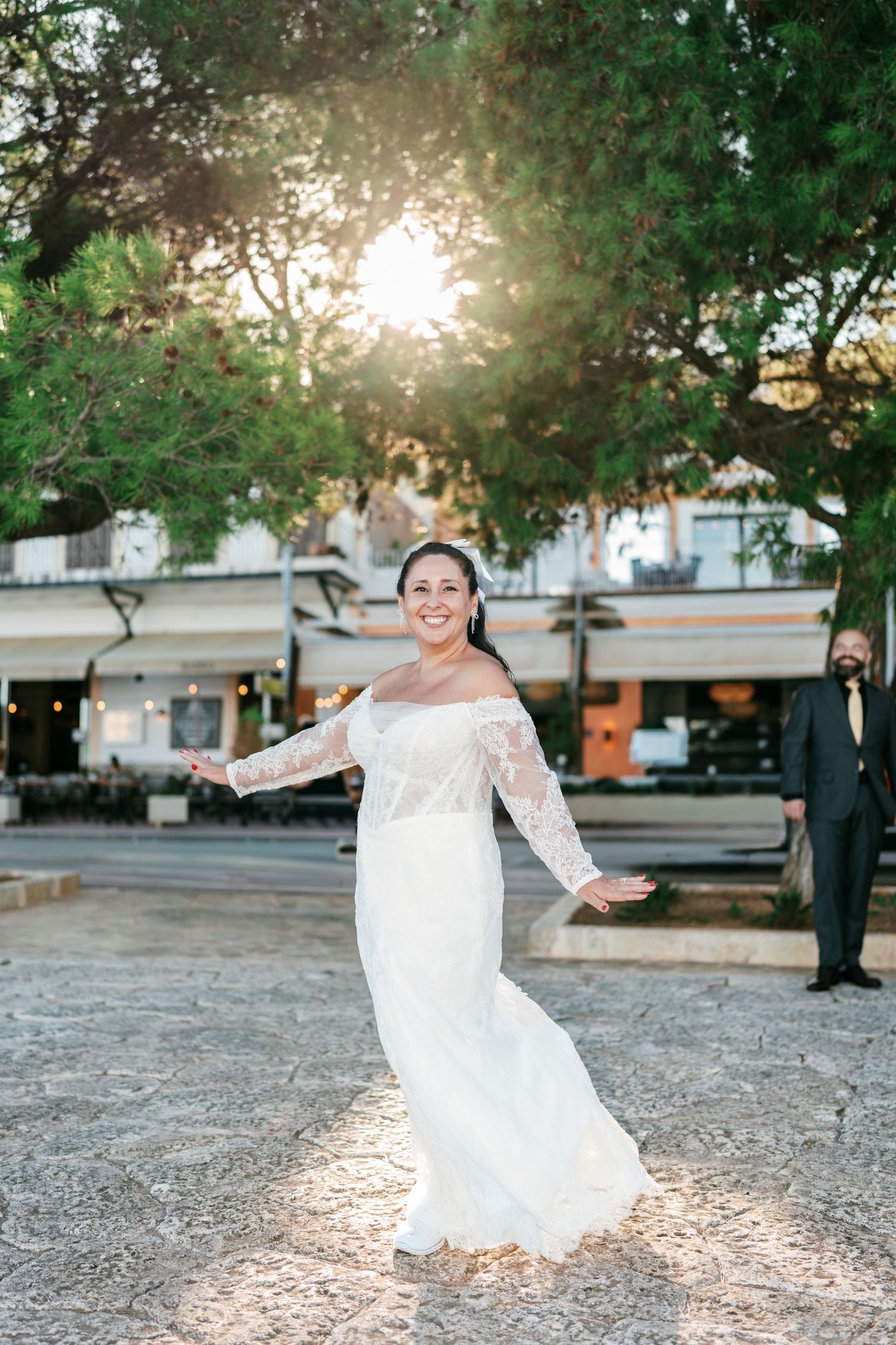 Smiling bride in a white lace gown twirls on a sunlit stone plaza while the groom in a dark suit watches from behind.