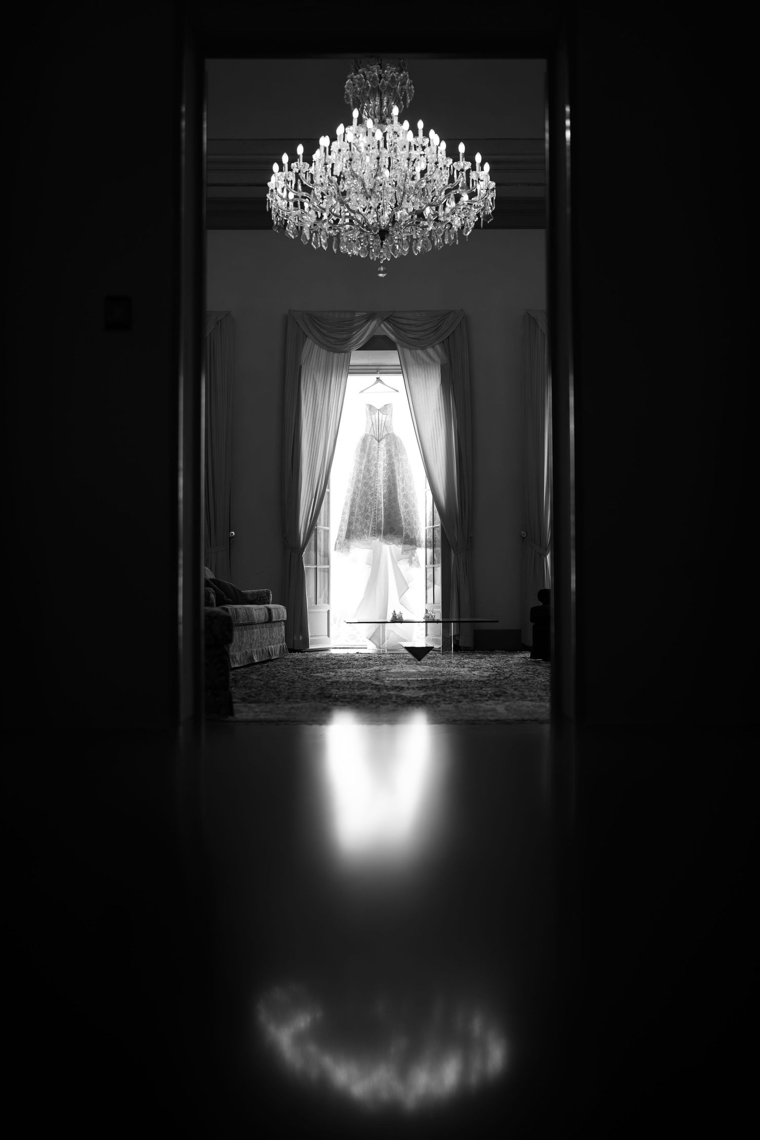 Black and white view through a doorway of a crystal chandelier and a wedding dress hanging by sheer curtains in an elegant room.