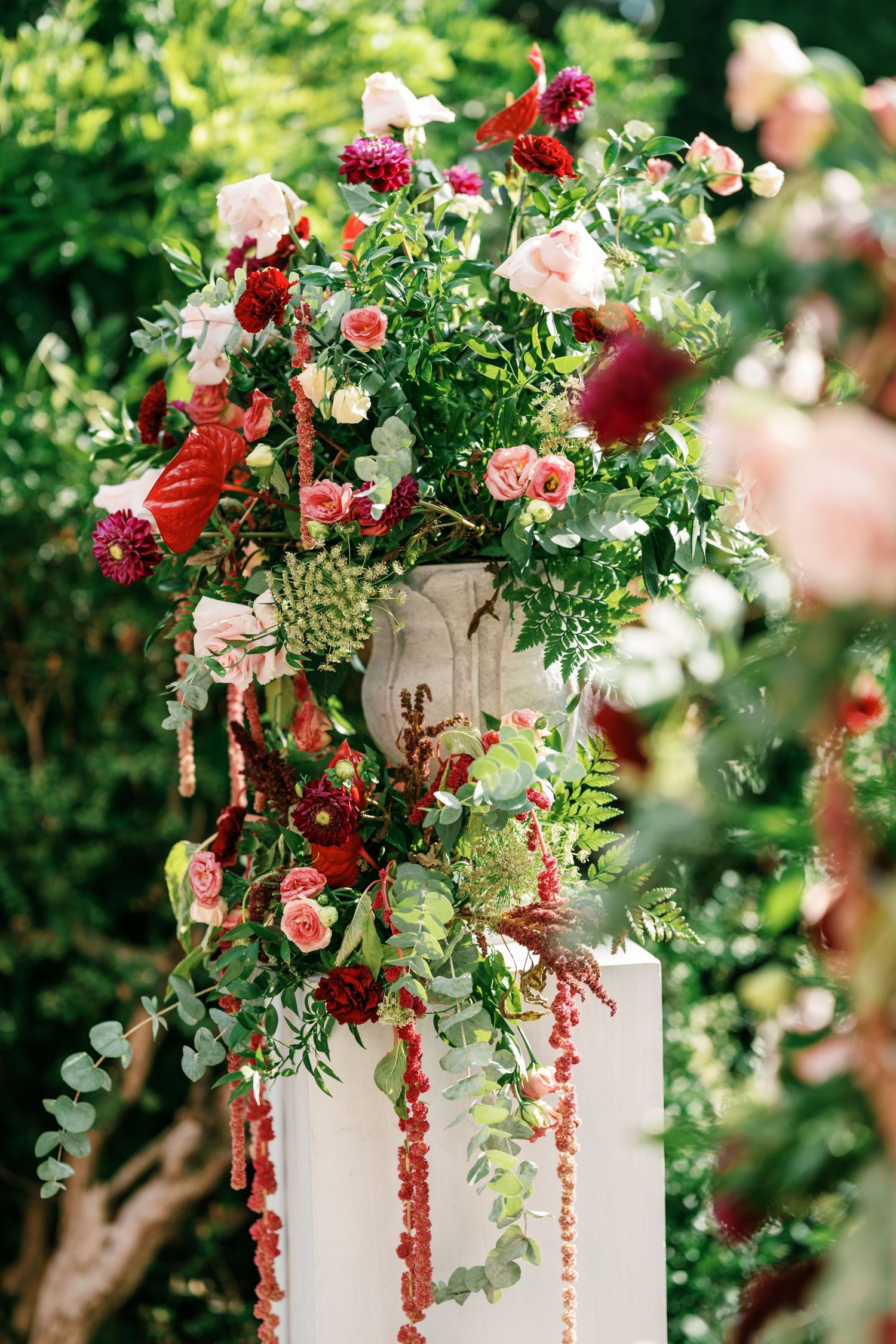 Lush wedding floral arrangement on a white pedestal with red and pink roses, dahlias, eucalyptus and trailing amaranthus in a garden.