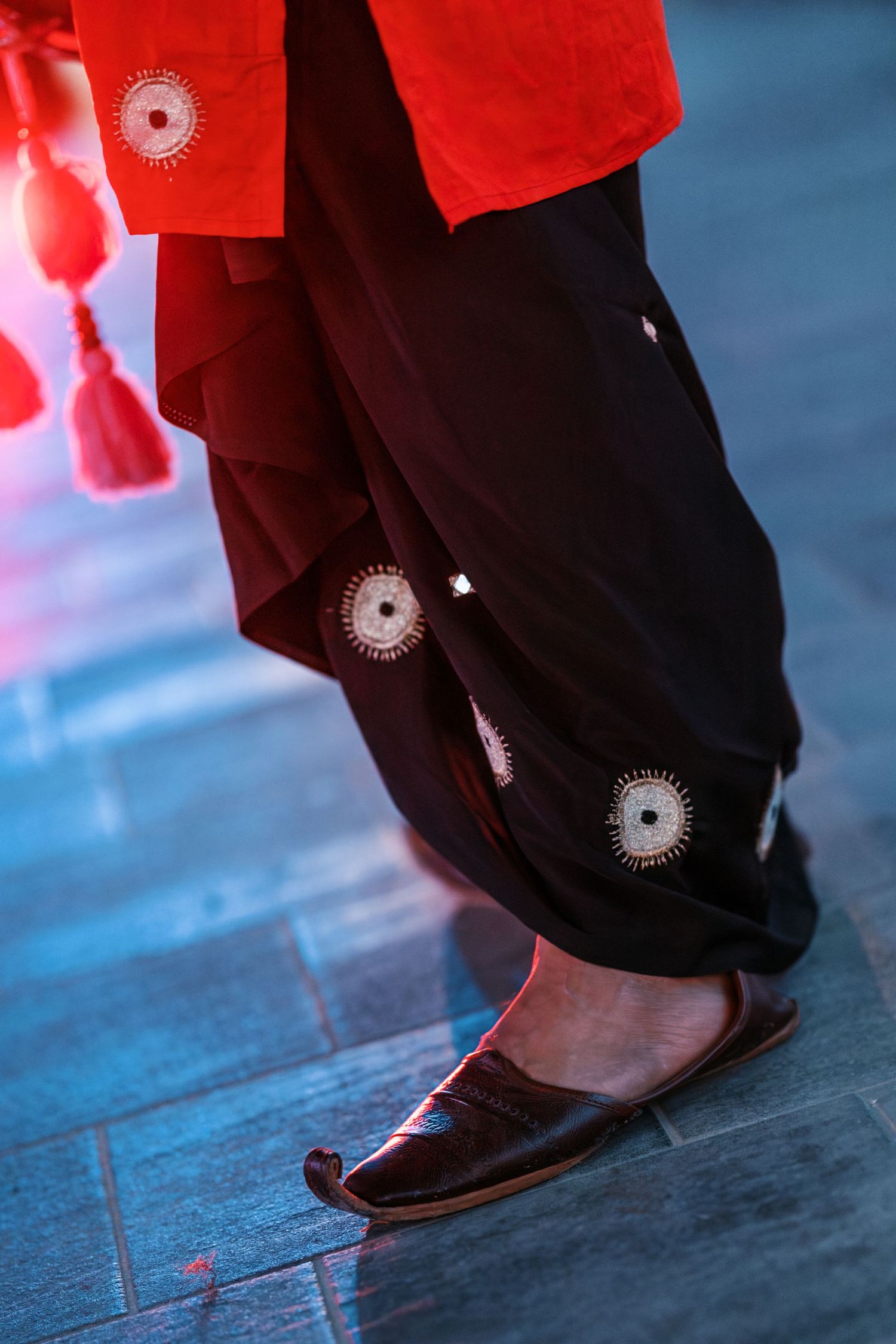 Close-up of a dancer's red kurta, black embroidered shalwar and curled leather mojari shoes beside a red tassel on a blue tiled floor.