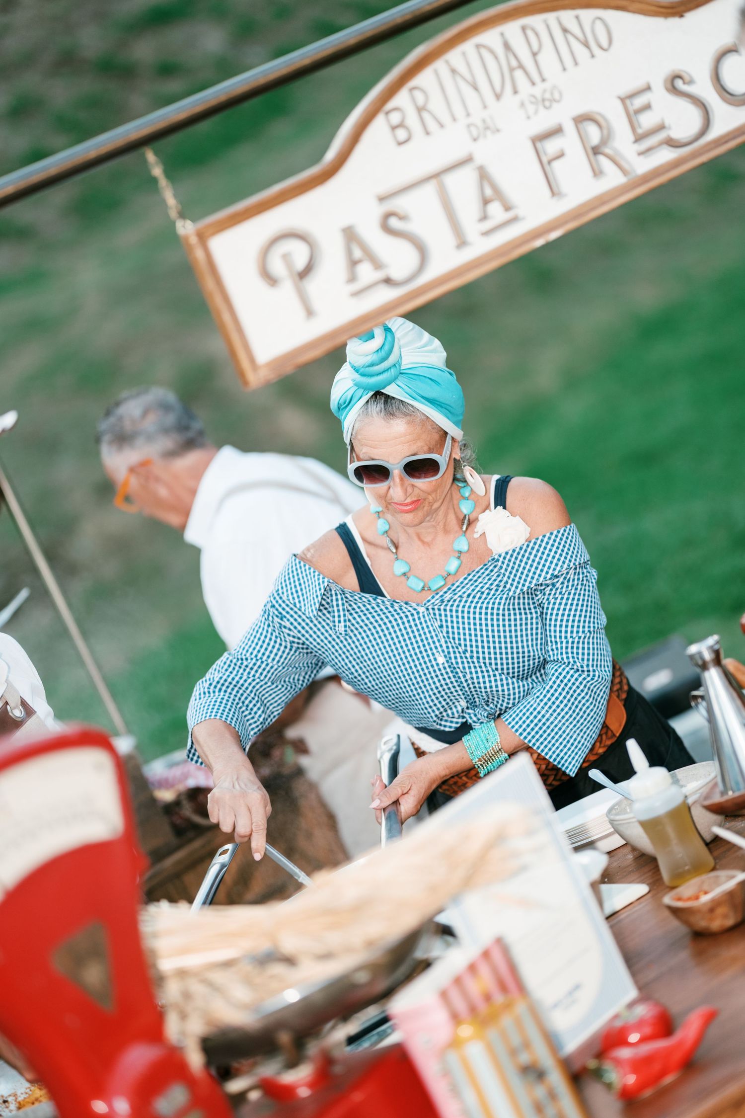 Woman in a blue gingham top, turquoise headscarf and sunglasses prepares fresh pasta at a rustic Pasta Fresca market stall.