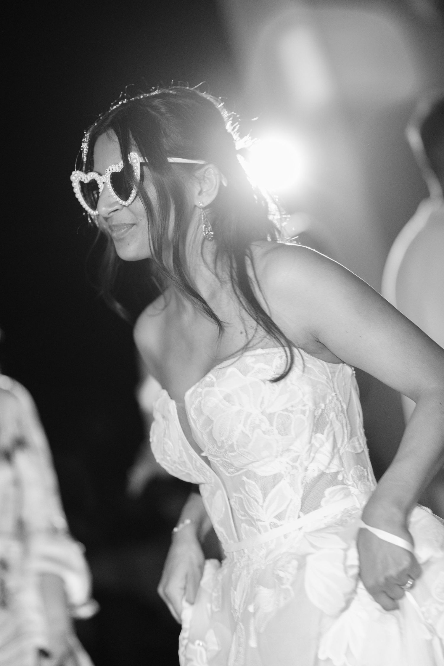 Black and white photo of a bride in a lace strapless wedding gown and heart-shaped sunglasses dancing at an evening reception.