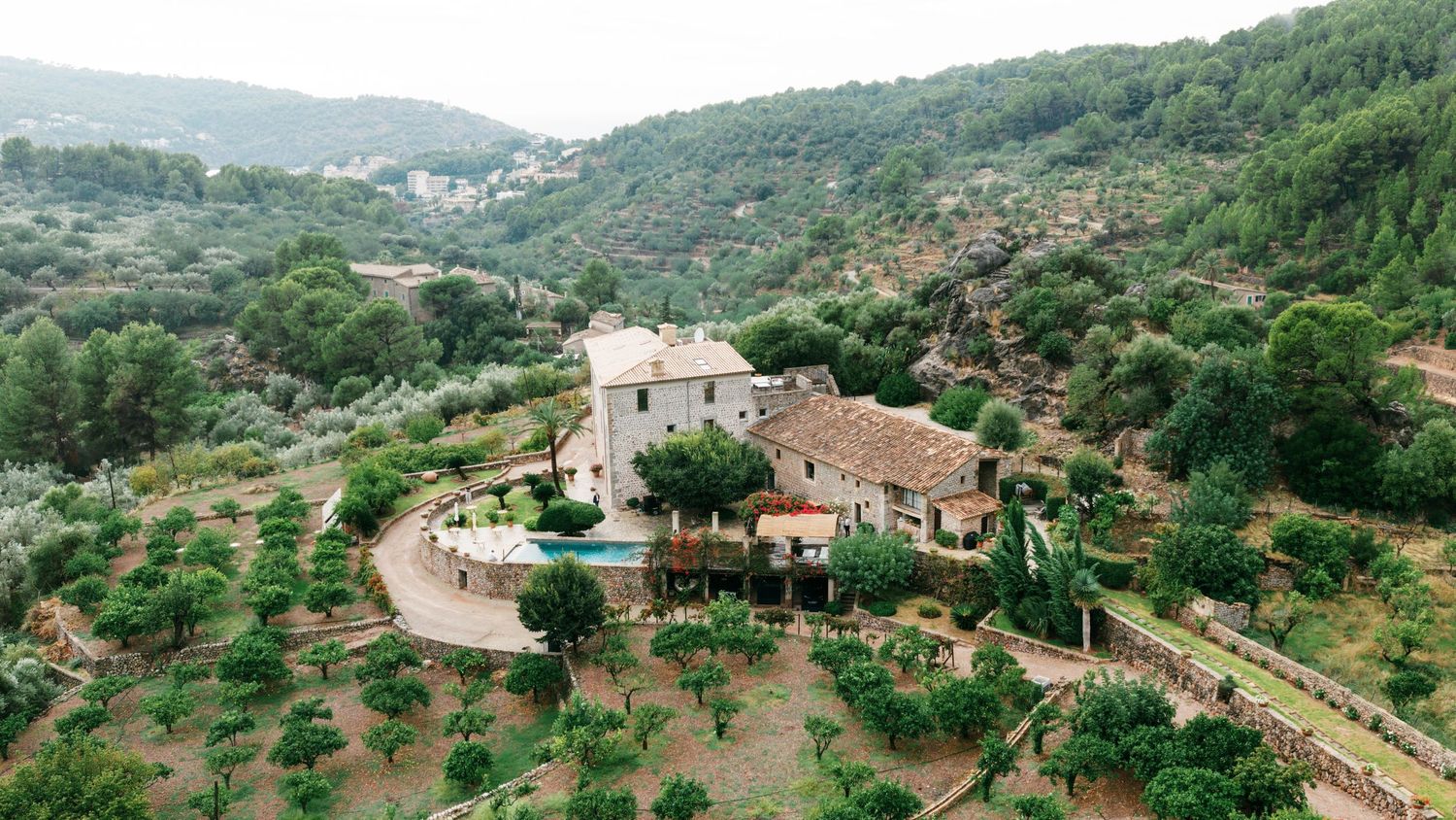 Aerial view of a stone countryside villa with terracotta roofs, a pool and terraced olive groves set among forested Mediterranean hills.
