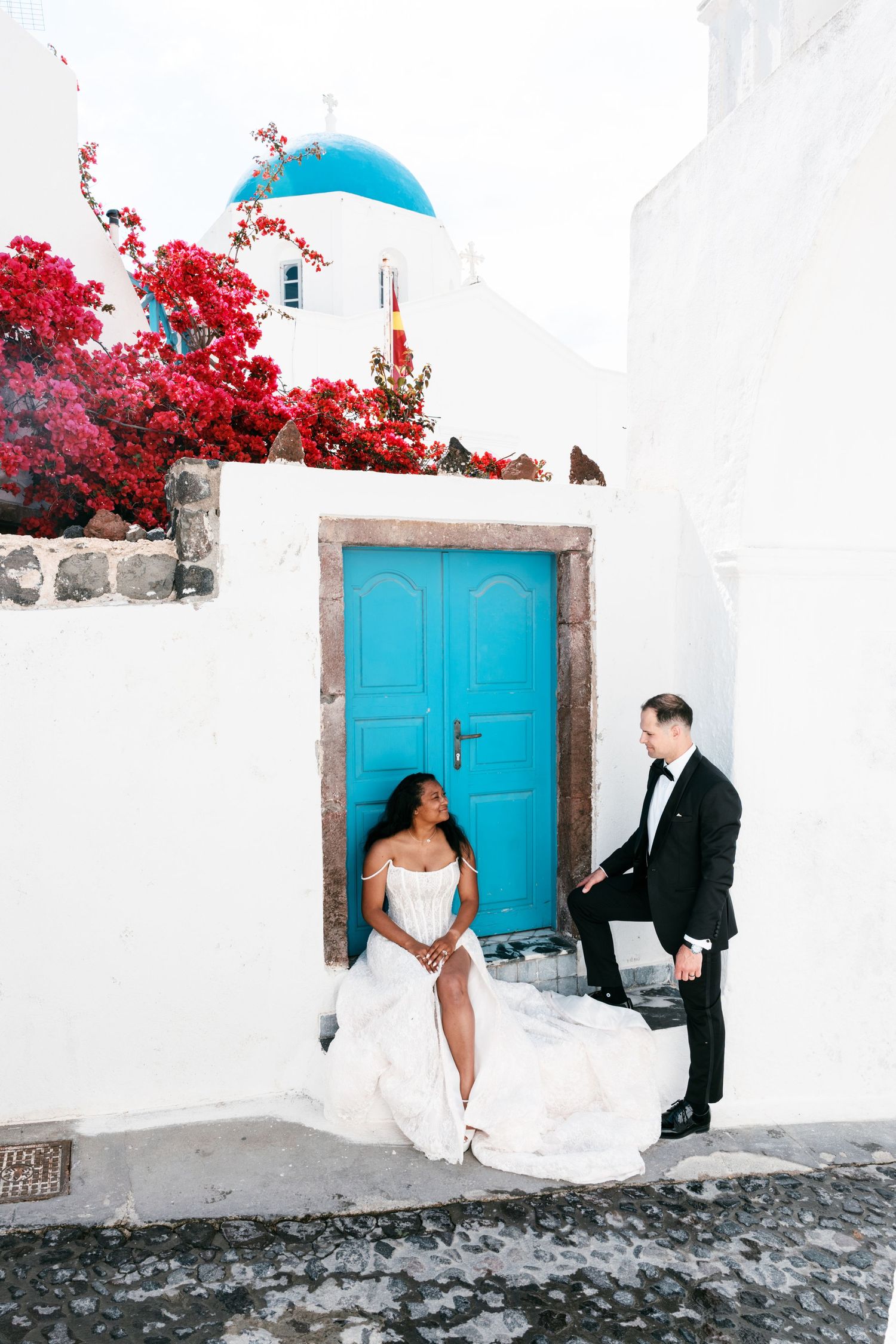 Interracial wedding couple poses by blue door in Santorini, Greece, with white walls and red bougainvillea blooms.
