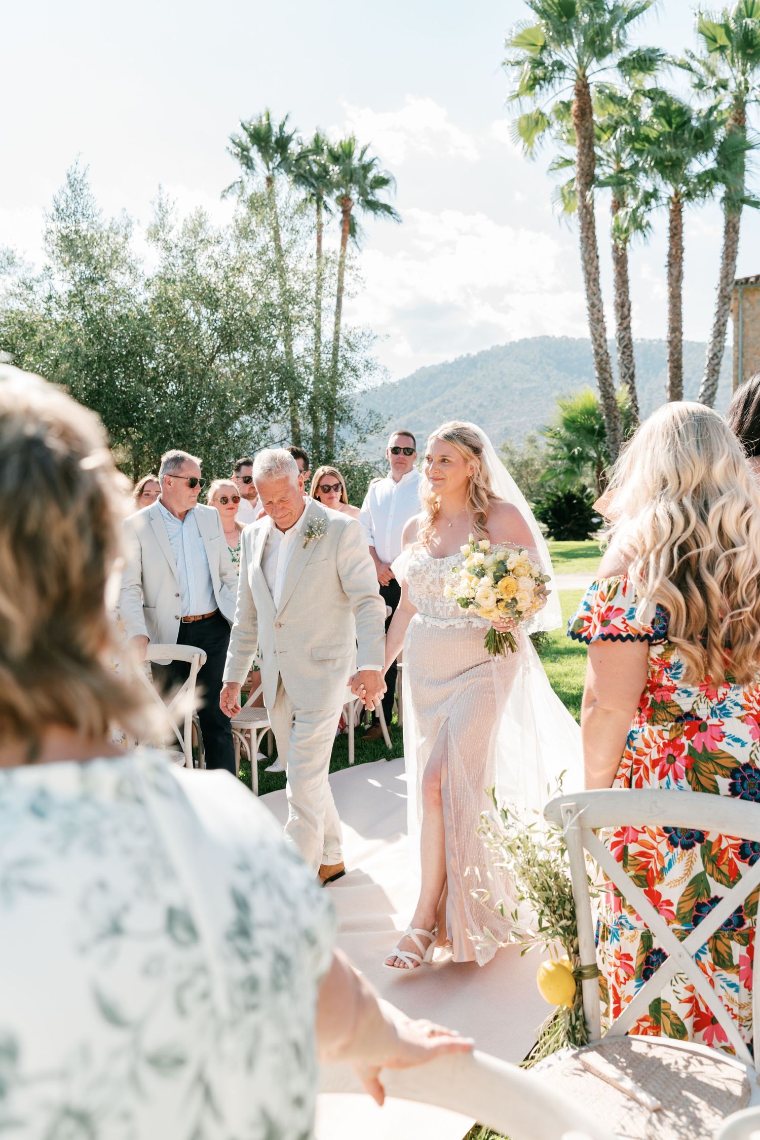 Bride walks down outdoor aisle with escort, holding yellow bouquet, palm trees and mountains in background. Mallorca wedding photographer