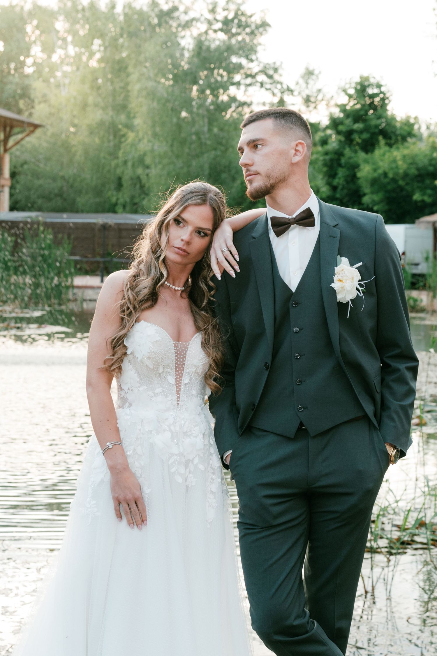 Elegant bride rests arm on grooms shoulder as couple poses by sunlit lakeside venue. Mallorca wedding photographer and filmmaker.
