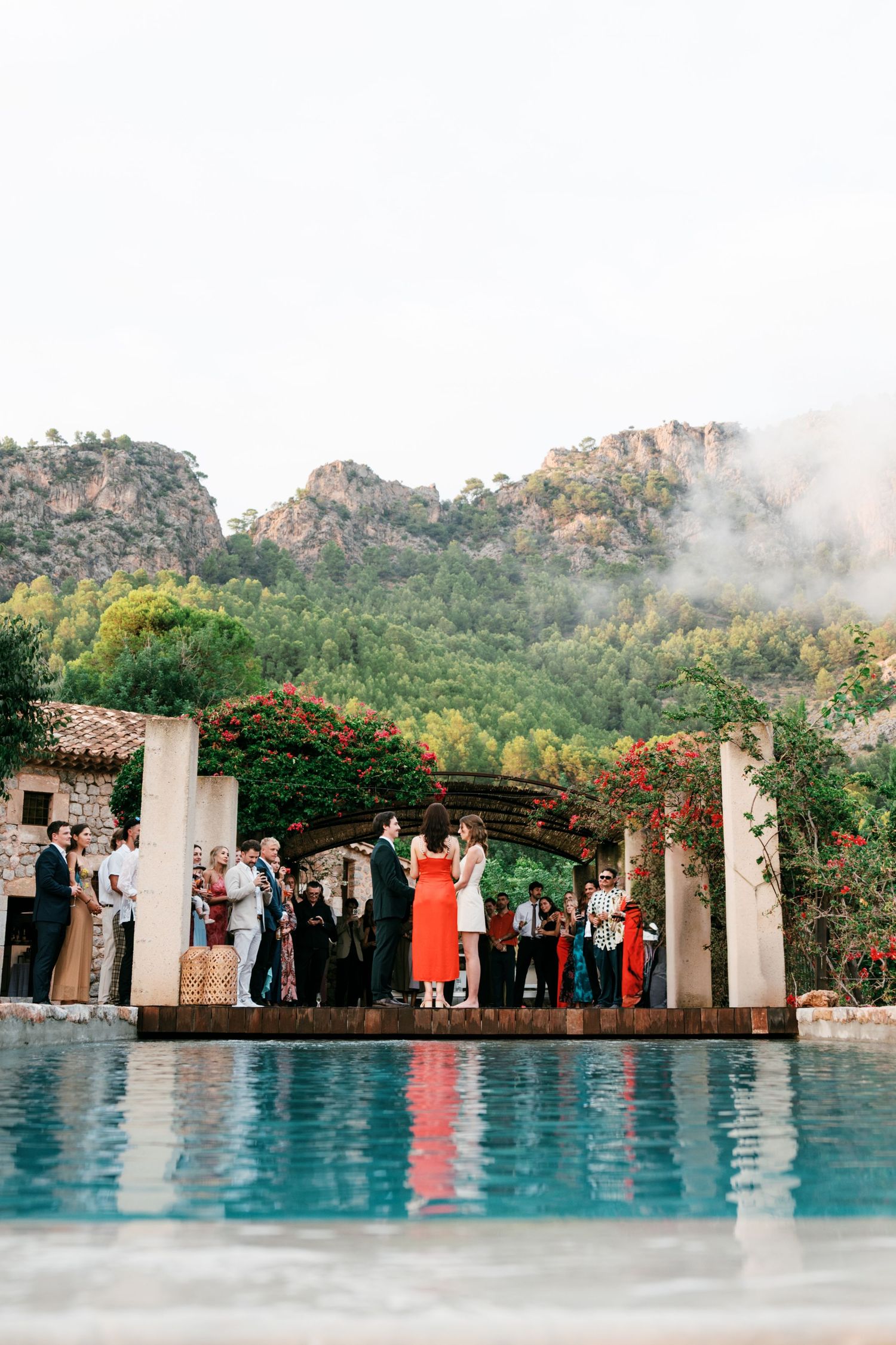 Wedding ceremony beside a turquoise pool with guests gathered under a flower-covered stone arch and forested mountains in the background.