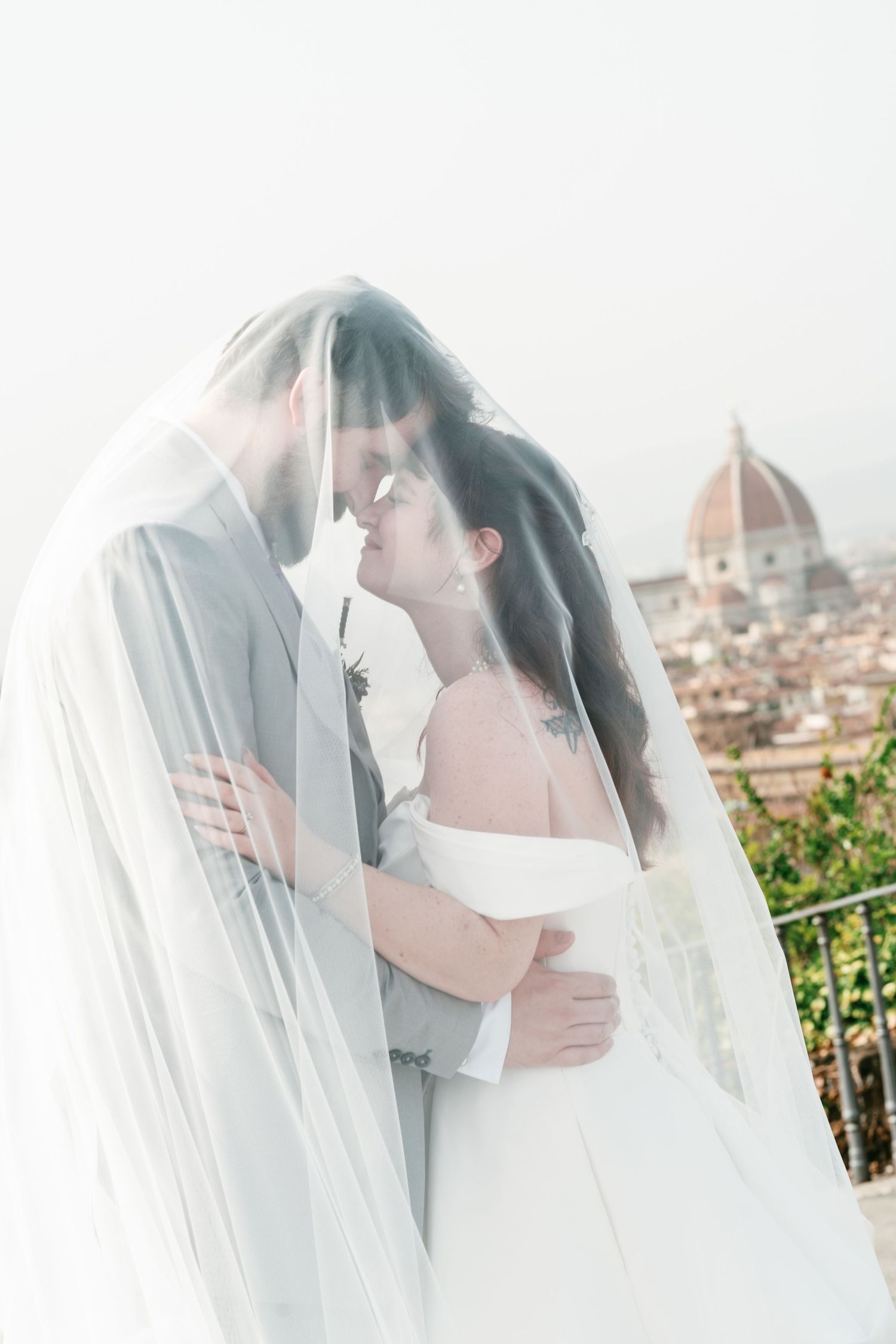 Bride and groom embrace forehead to forehead beneath a sheer veil, with the Florence Cathedral dome rising over the city behind them.