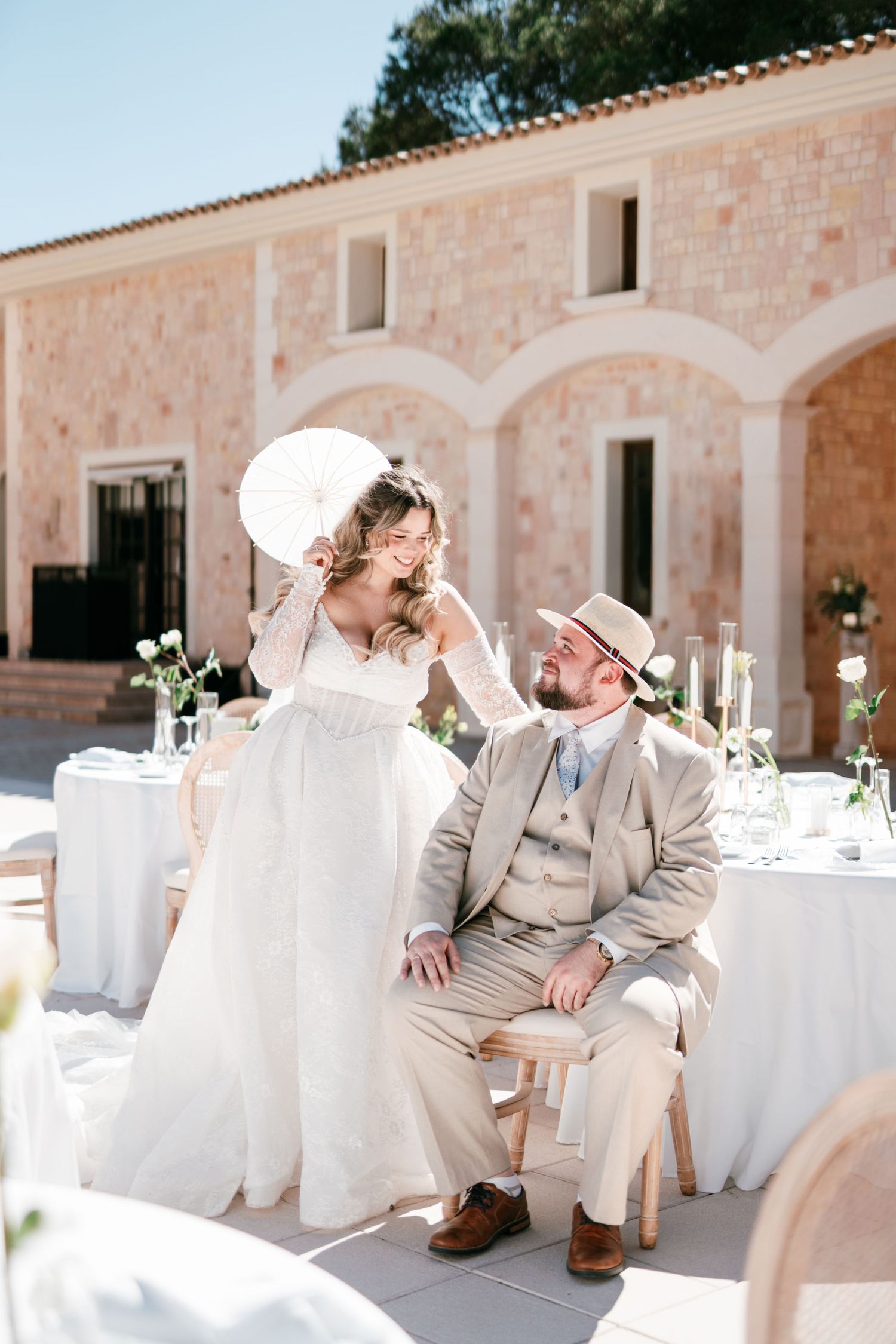 Bride in white gown with parasol and groom in tan suit share a joyful moment at outdoor wedding reception in Mallorca.