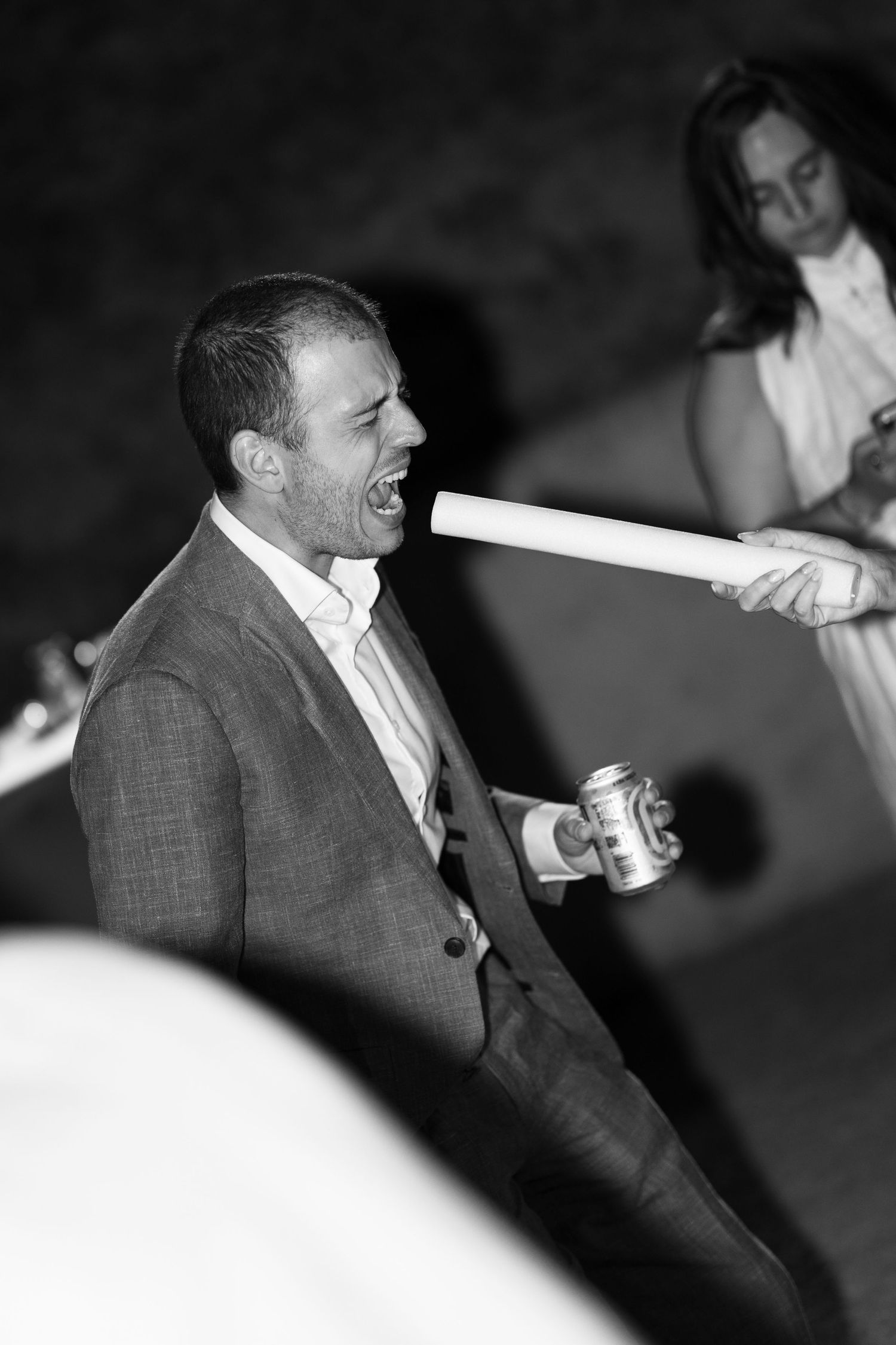 Black and white photo of a man in a suit laughing while someone points a tube at his face at a party.