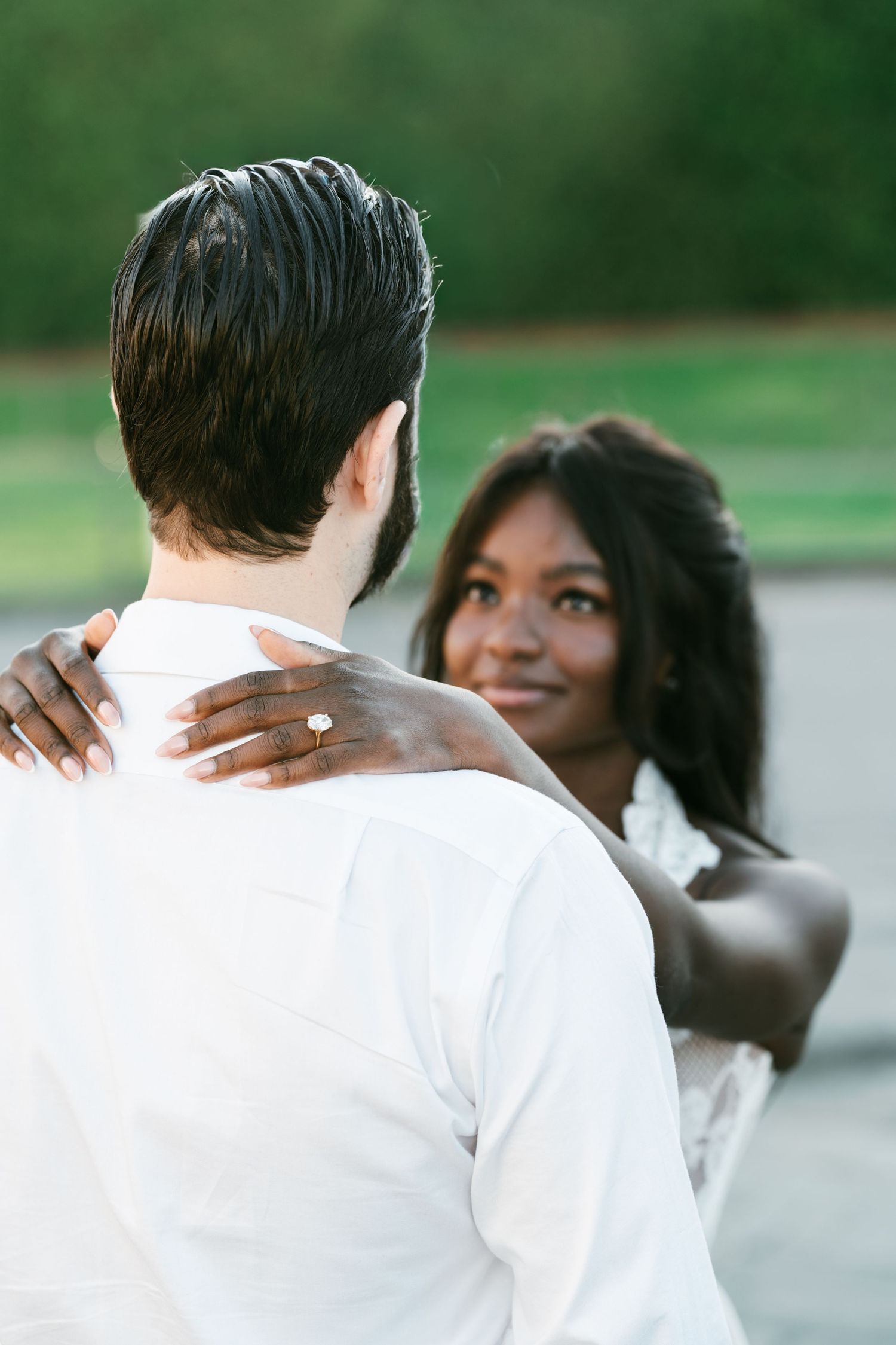 A woman with a sparkling diamond engagement ring smiles softly as she embraces a bearded man in a white shirt outdoors.