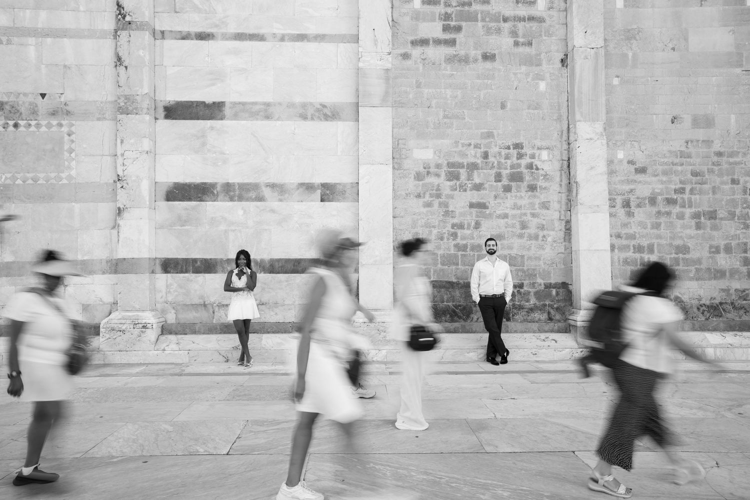 Black and white photo of motion-blurred pedestrians walking past two still figures standing against an old stone brick wall.