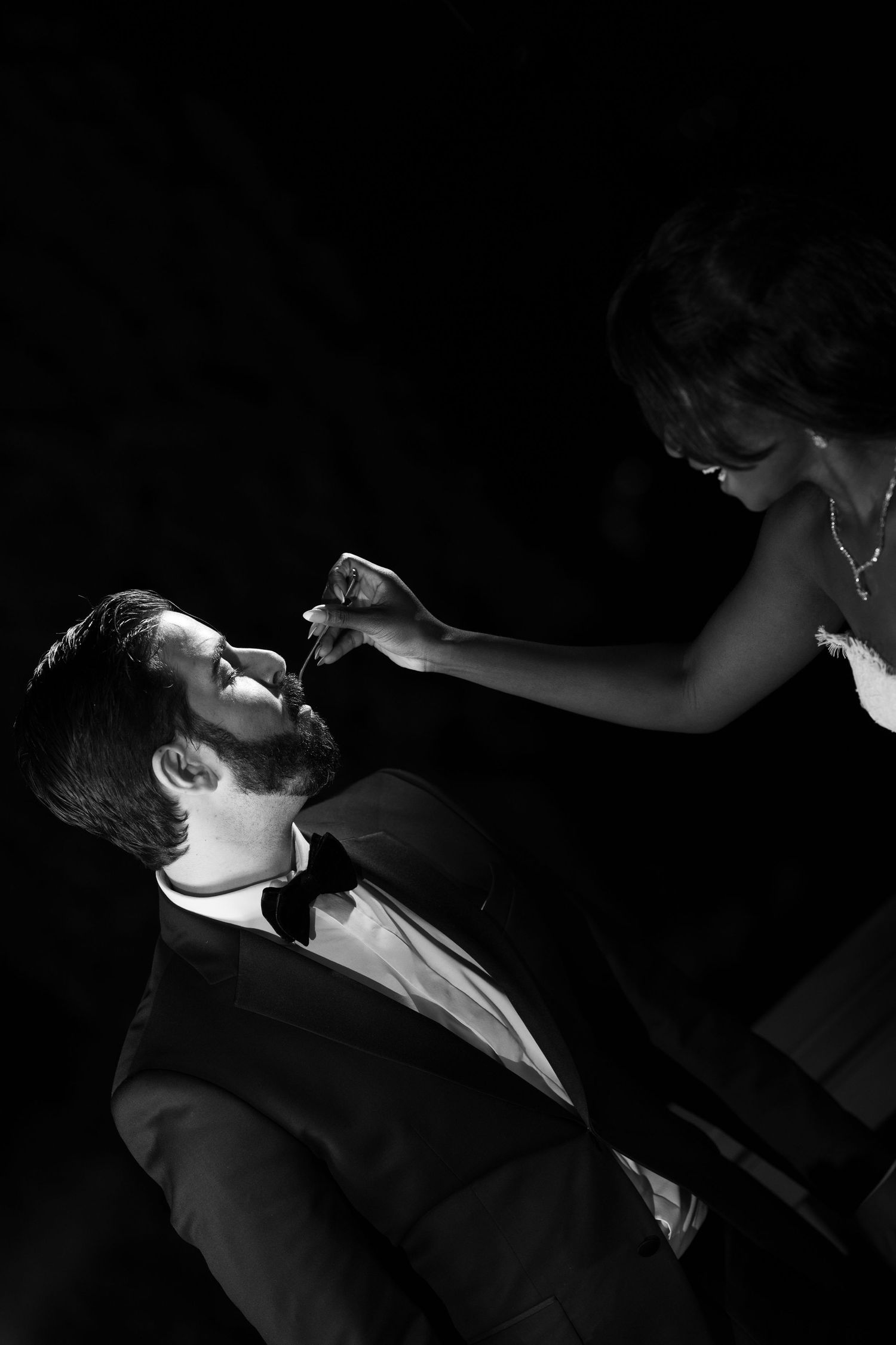 Black and white photo of a bride in a necklace feeding a bearded groom in a tuxedo and bow tie against a dark background.