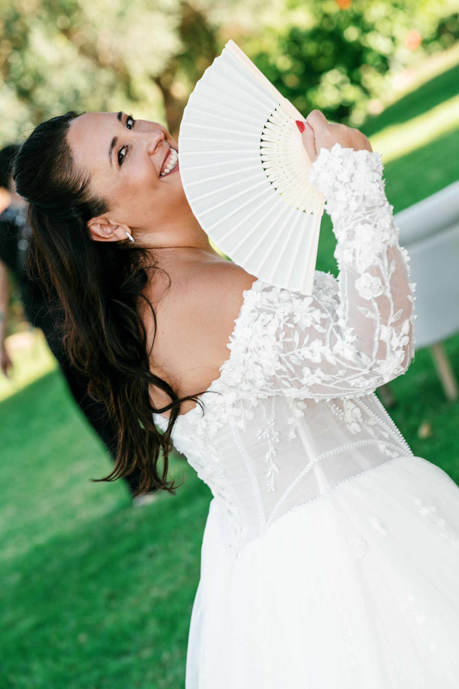 Smiling bride in an off-shoulder white lace wedding gown holds a white folding fan beside her face on a sunny green lawn.