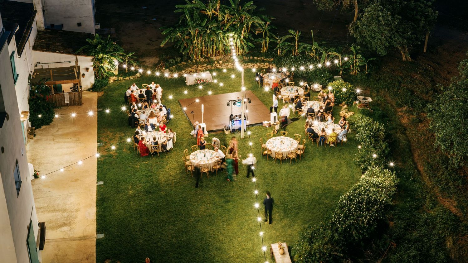 Aerial view of an outdoor garden wedding reception with round tables, guests, and glowing string lights at dusk.