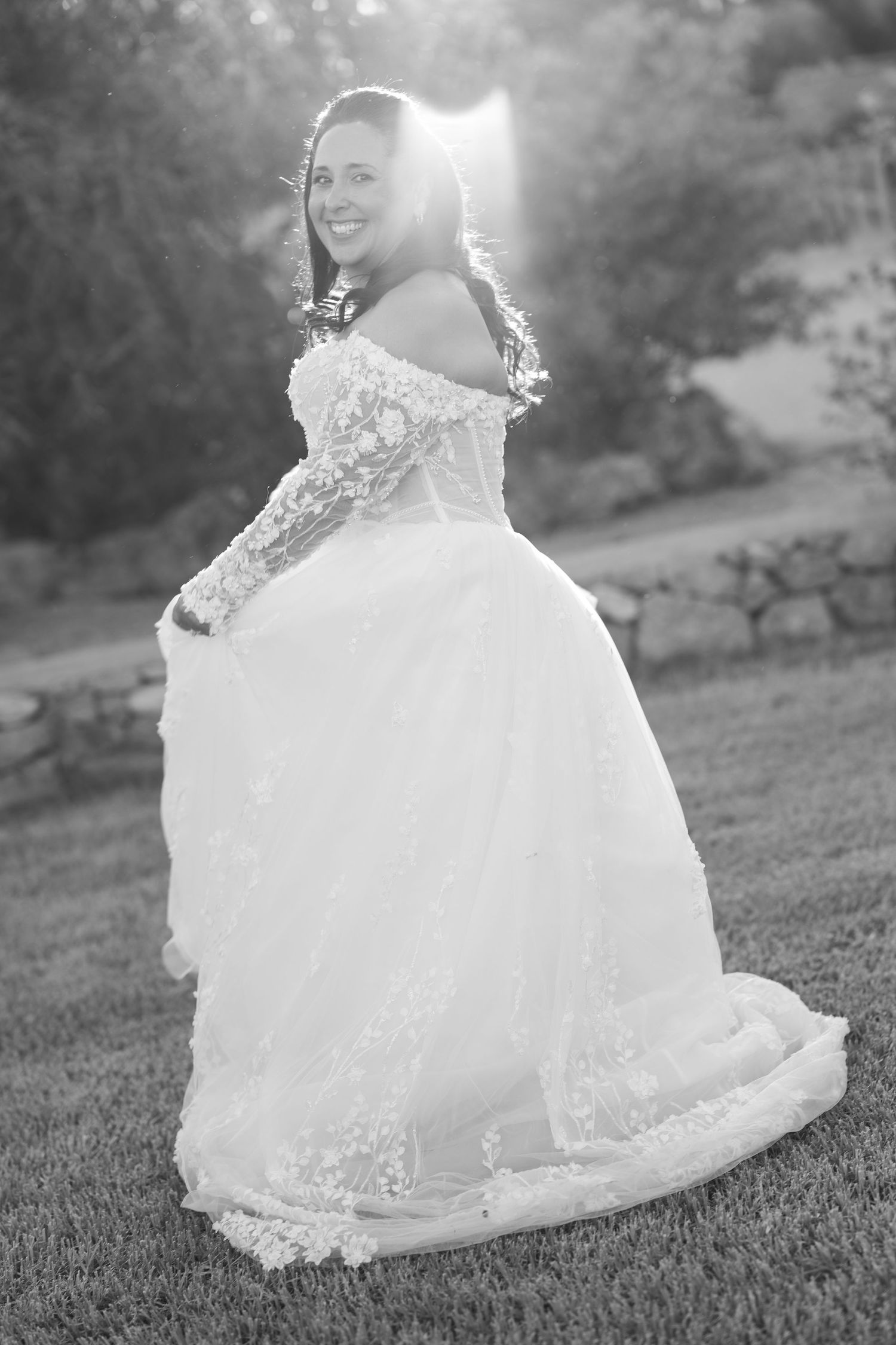 Smiling bride in an off-shoulder lace wedding gown with a full tulle skirt twirls on a grassy lawn in glowing black and white sunlight.