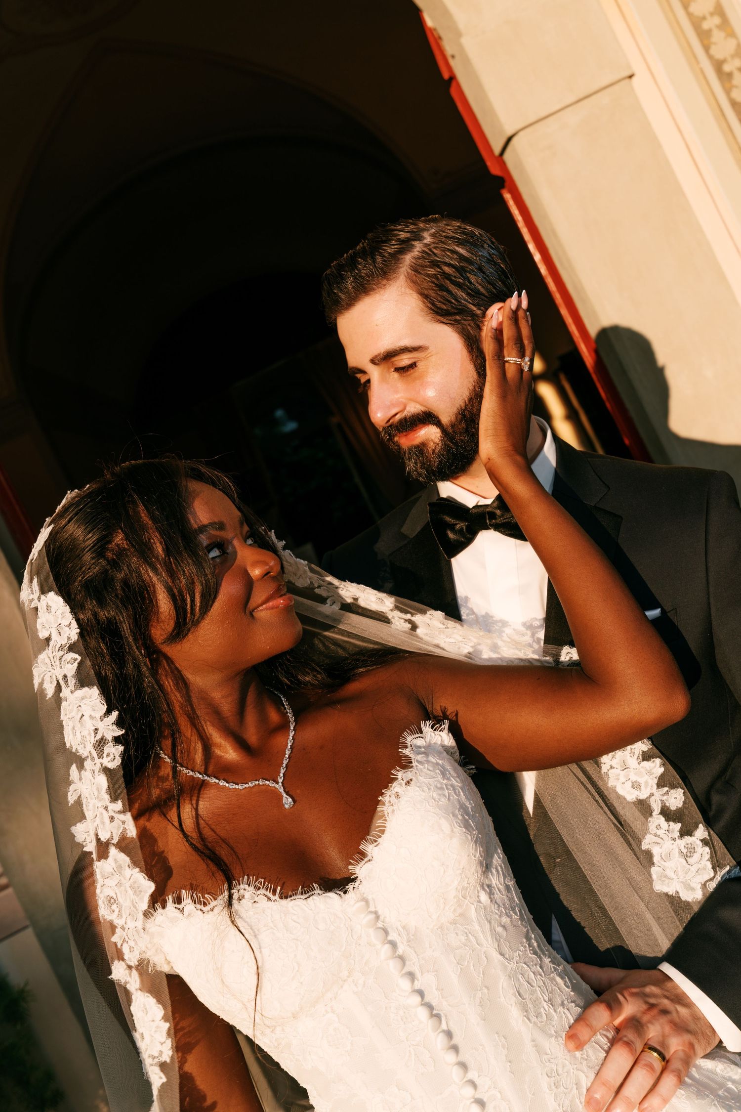 Smiling bride in a lace wedding gown and veil touches her hair as her bearded groom in a black tuxedo and bow tie gazes at her.