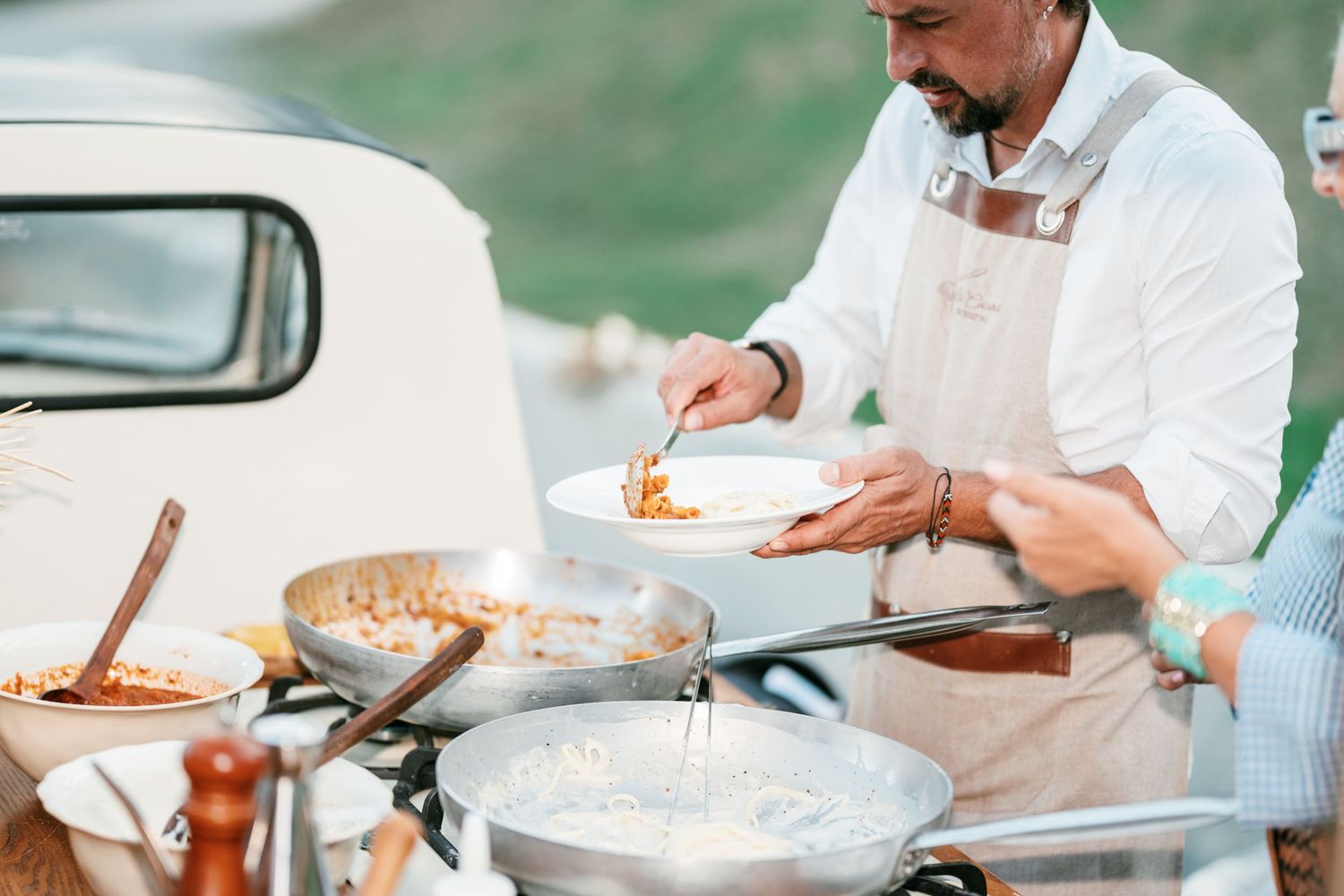 Chef in a white shirt and apron serves freshly cooked pasta from large pans at an outdoor catering event beside a vintage van.