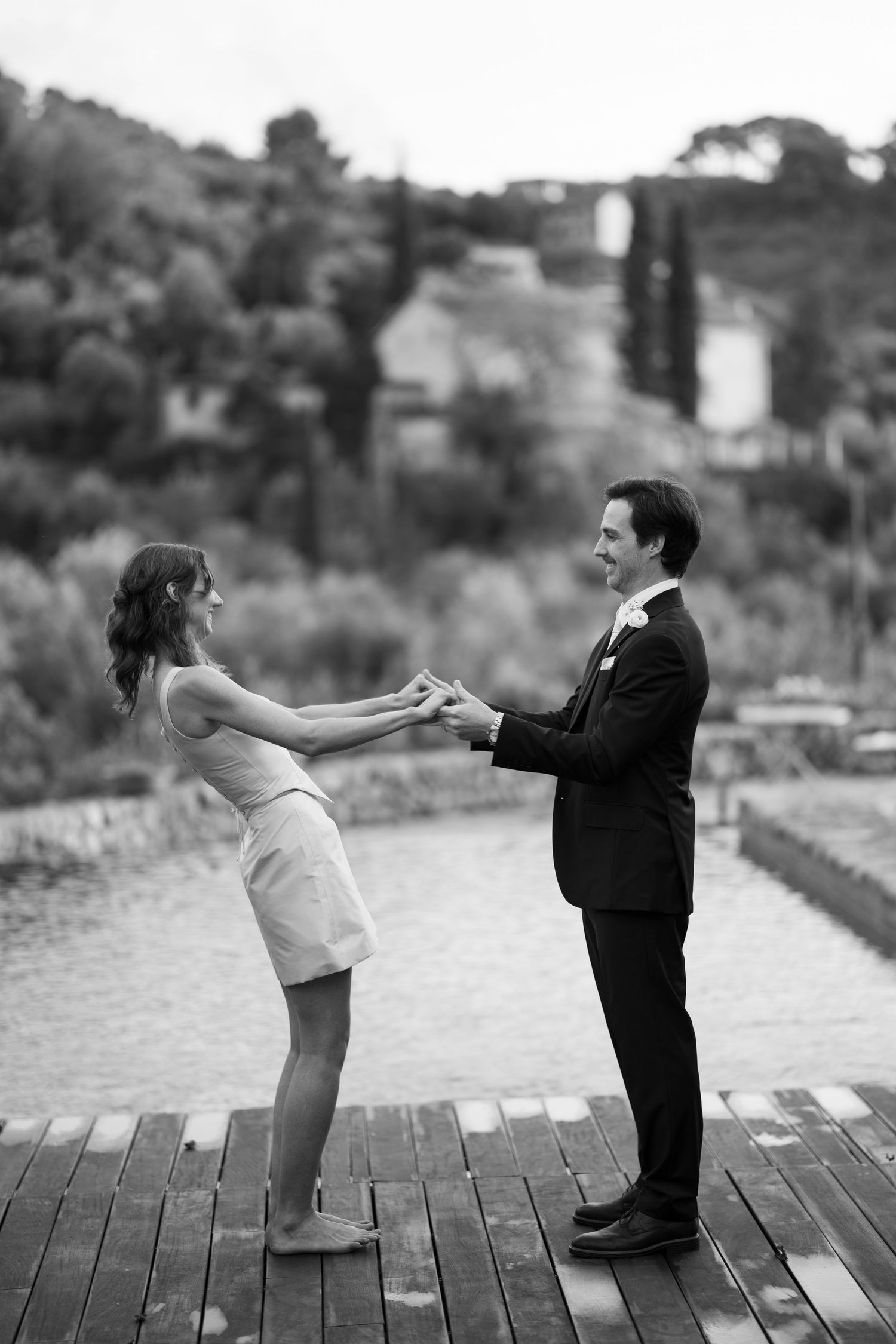 Black and white photo of a bride and groom holding hands on a wooden dock facing each other beside a calm lake.