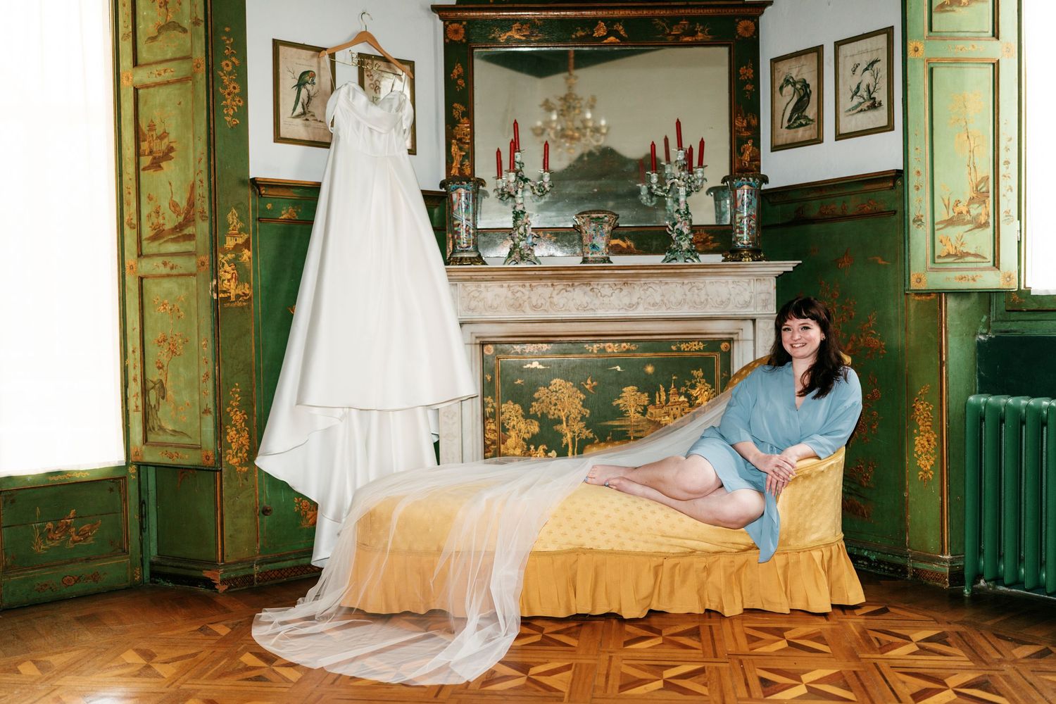Bride in a blue robe reclines on a gold daybed beside her white wedding dress hanging in an ornate green room with a fireplace.