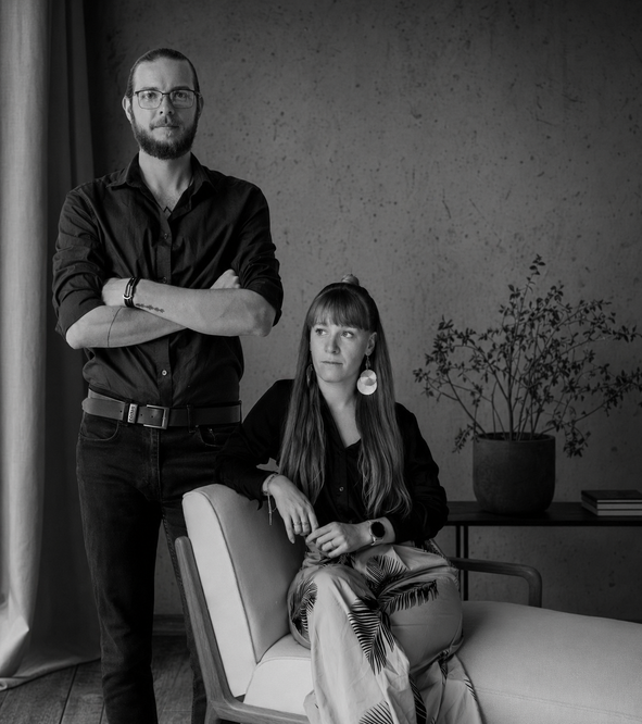 Black and white portrait of a man and woman in modern interior, man standing with crossed arms, woman seated in chair.