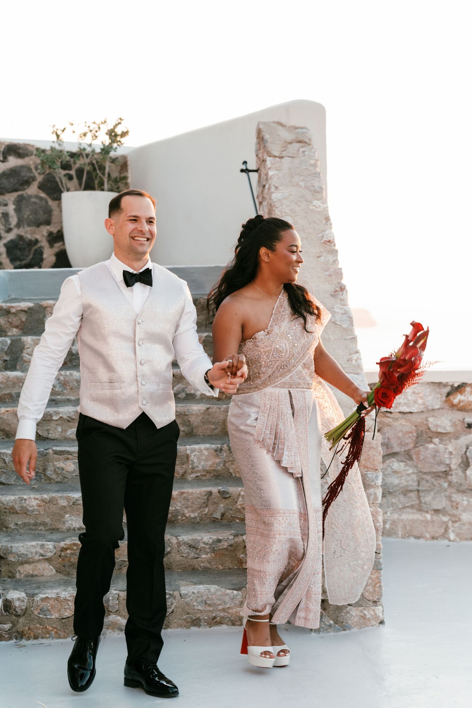 Smiling couple in formal attire walk hand-in-hand, woman holding red flowers, at an outdoor Mediterranean venue.