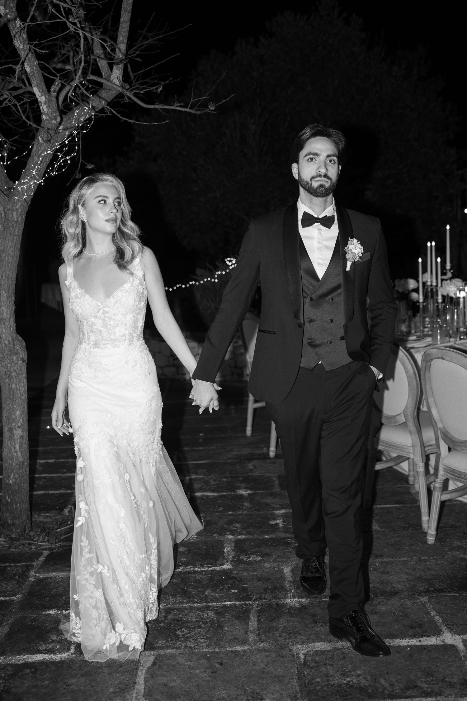 Black and white photo of bride and groom walking hand in hand at candlelit outdoor reception. Puglia wedding photographer, film