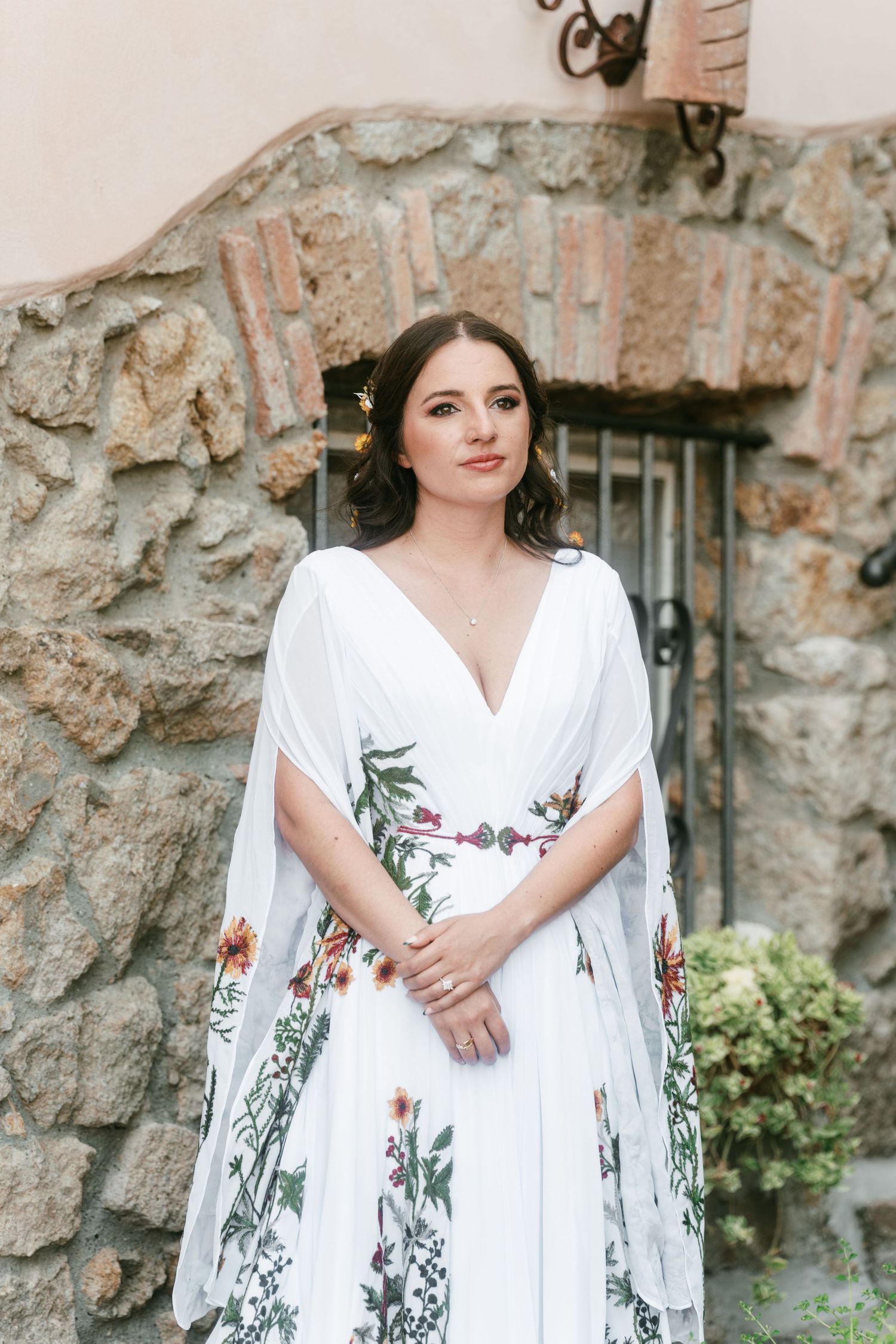 Bride in a stunning white floral embroidered gown with cape sleeves standing before a stone wall. Tuscany wedding photographer, filmmaker