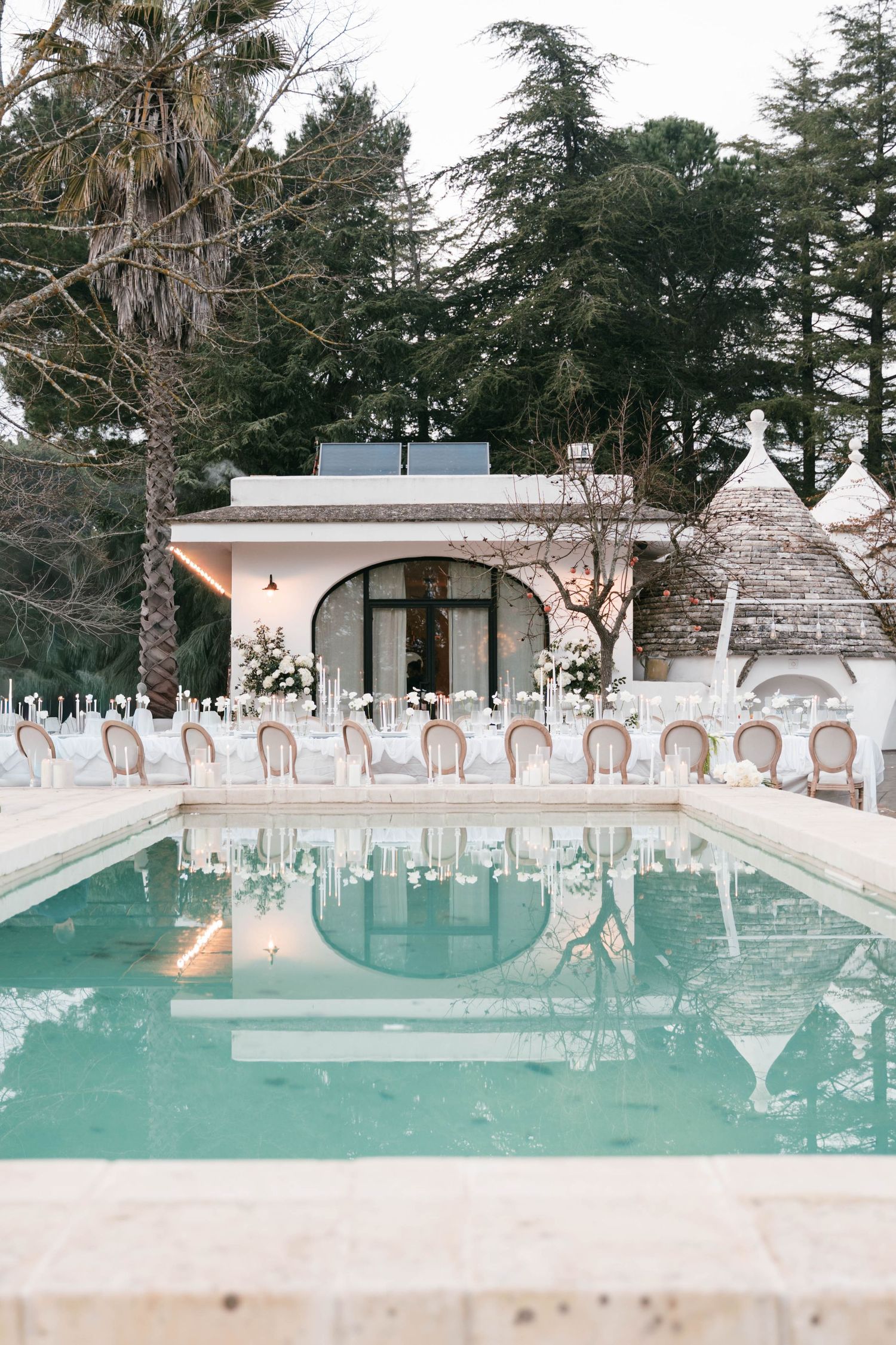 Elegant outdoor wedding reception setup beside reflective pool with white floral decorations and trullo. Puglia wedding photographer,