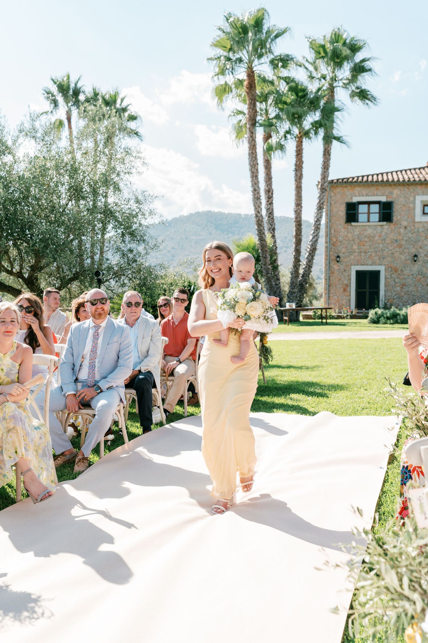 Woman in gold dress carries baby down outdoor wedding aisle, guests seated, palm trees and stone villa in background. Mallorca wedding photo