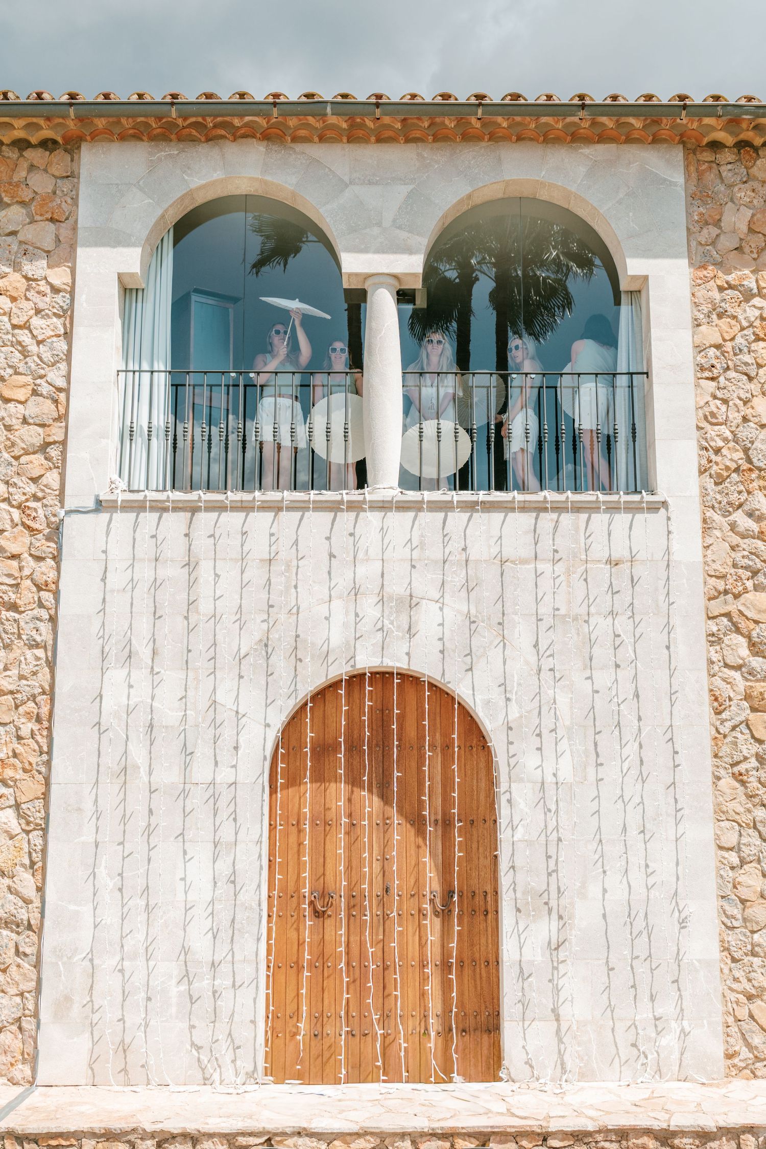 People standing on a balcony with arched windows of a stone building with a wooden arched door below. Mallorca wedding filmmaker, video