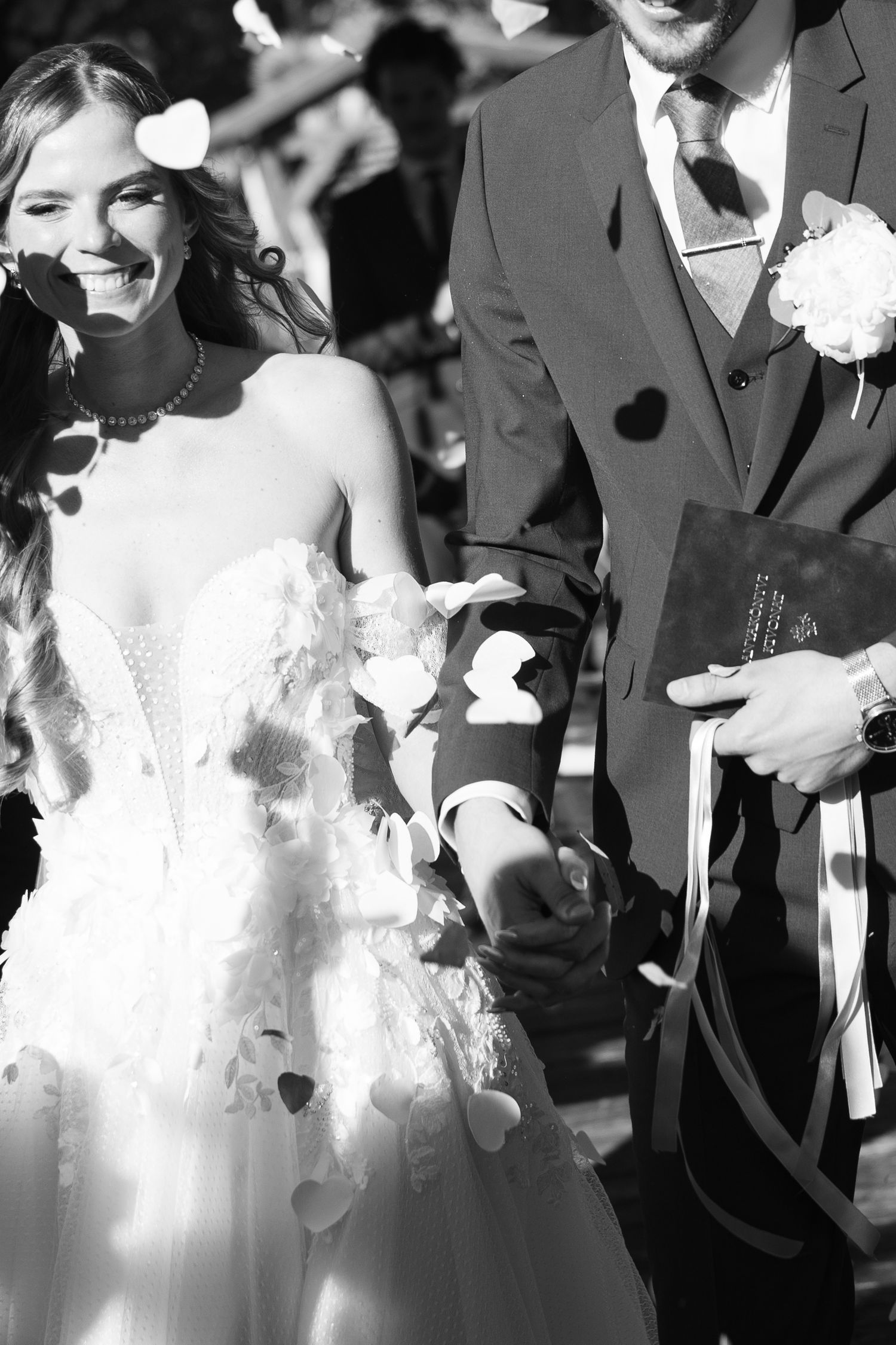 Black and white photo of radiant bride and groom walking through heart-shaped confetti shower. Mallorca wedding photographer and filmmaker.