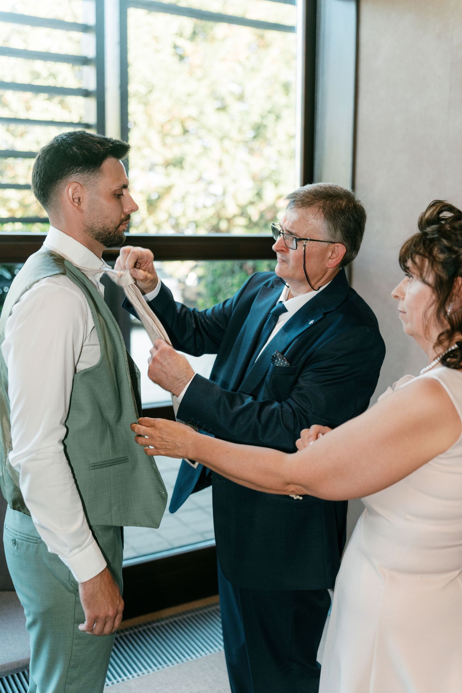 Older man adjusting a grooms tie while a woman assists during wedding preparations indoors. Italy wedding photographer and filmmaker