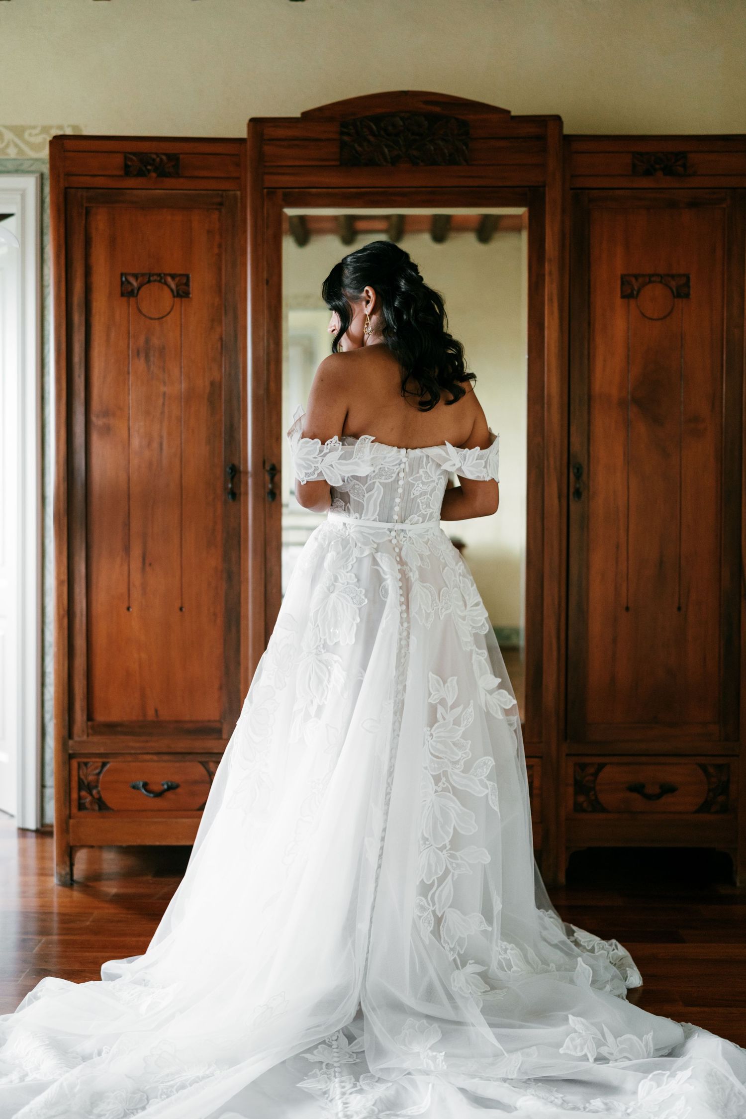 Bride in an off-shoulder lace wedding gown with a long flowing train stands facing a vintage wooden wardrobe with an open mirror.
