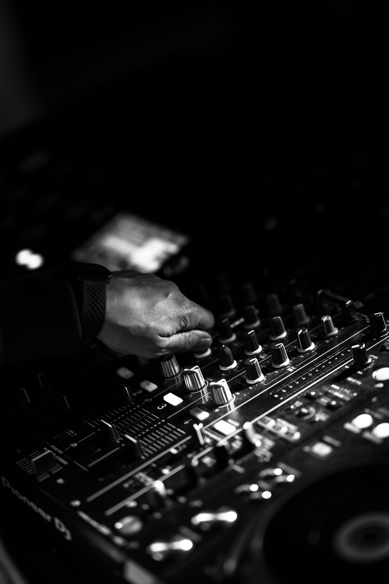 Black and white photo of a DJ's hand adjusting knobs on a professional mixing console and turntable in a dark club setting.