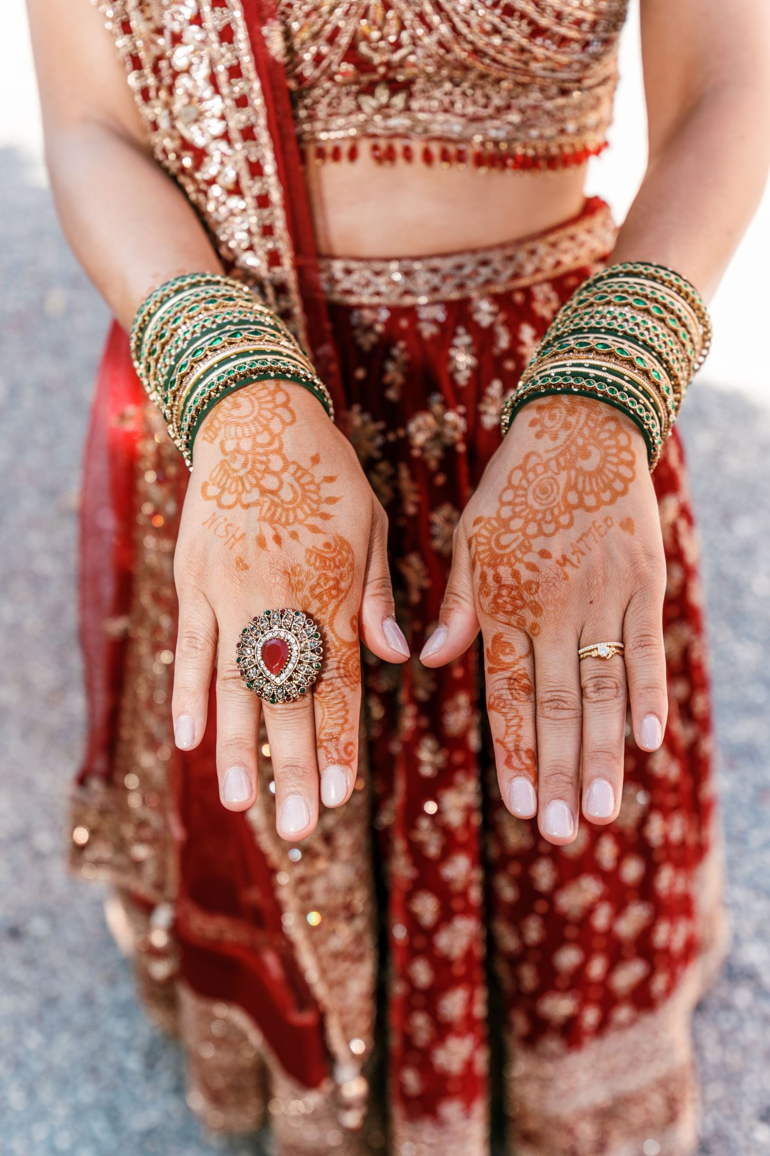 Bride's hands decorated with intricate henna, green bangles and a large red cocktail ring over a red embroidered lehenga.