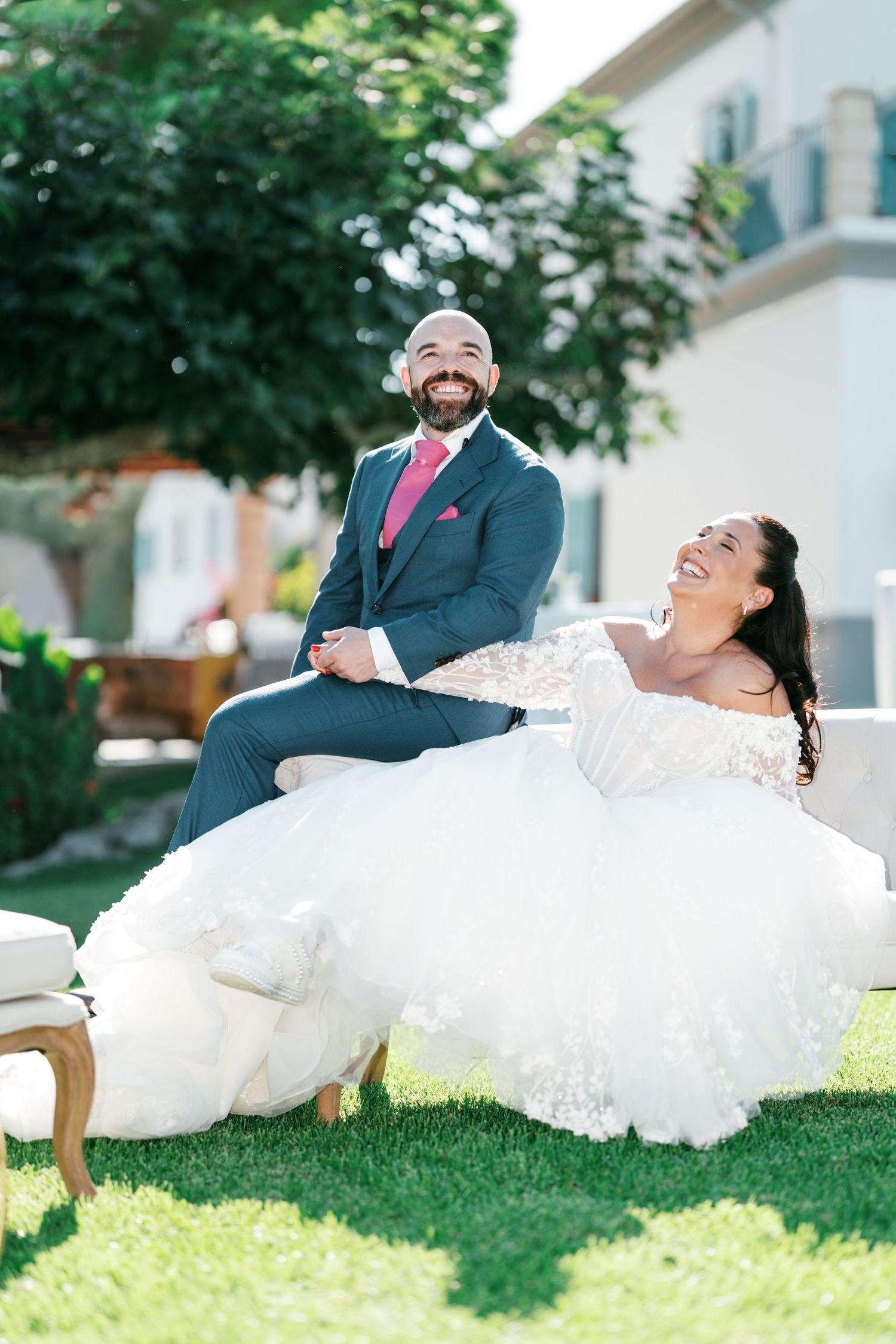 Smiling bride in an off-shoulder white lace gown and groom in a blue suit with pink tie laughing together on a sunny garden lawn.
