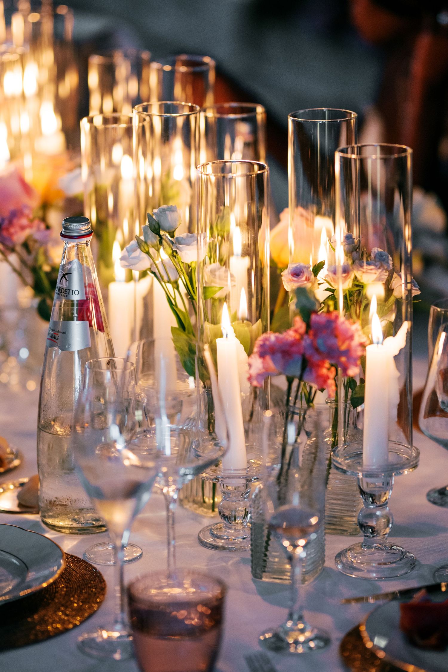 Wedding table centerpiece with lit white candles in glass cylinders, pink flowers, wine glasses and a water bottle at dusk.