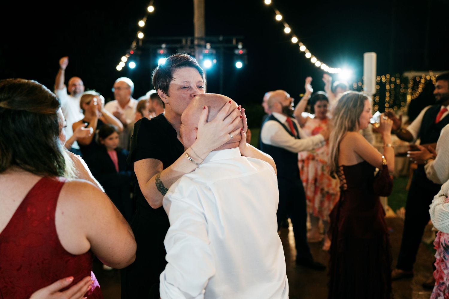 Wedding guests embrace and cheer on the dance floor under string lights as a bald man is hugged during a lively night reception.