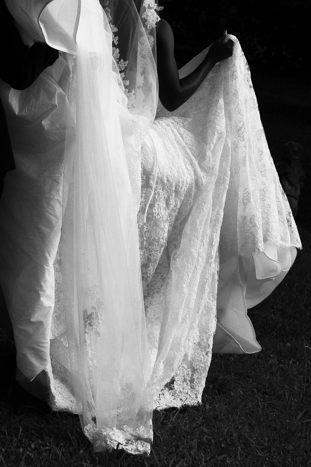 Black and white photo of a bride lifting the flowing lace skirt and long veil of her wedding gown while walking across the grass.