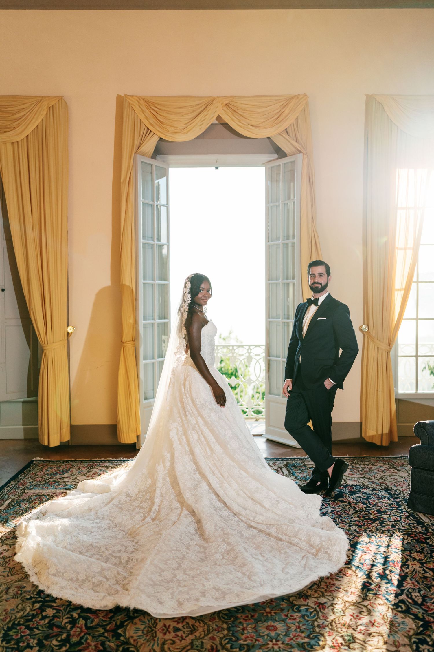 Bride in a lace gown with a long flowing train and groom in a black tuxedo pose by open balcony doors with gold drapes.