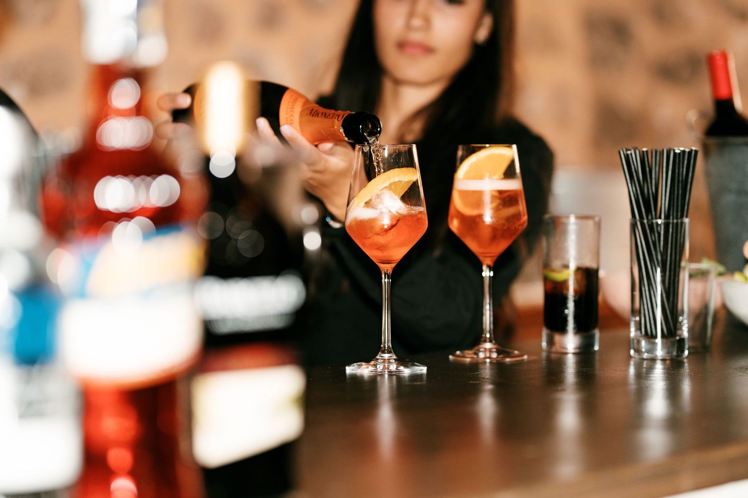 A bartender pours prosecco into two glasses of orange Aperol spritz garnished with orange slices at a bar counter.