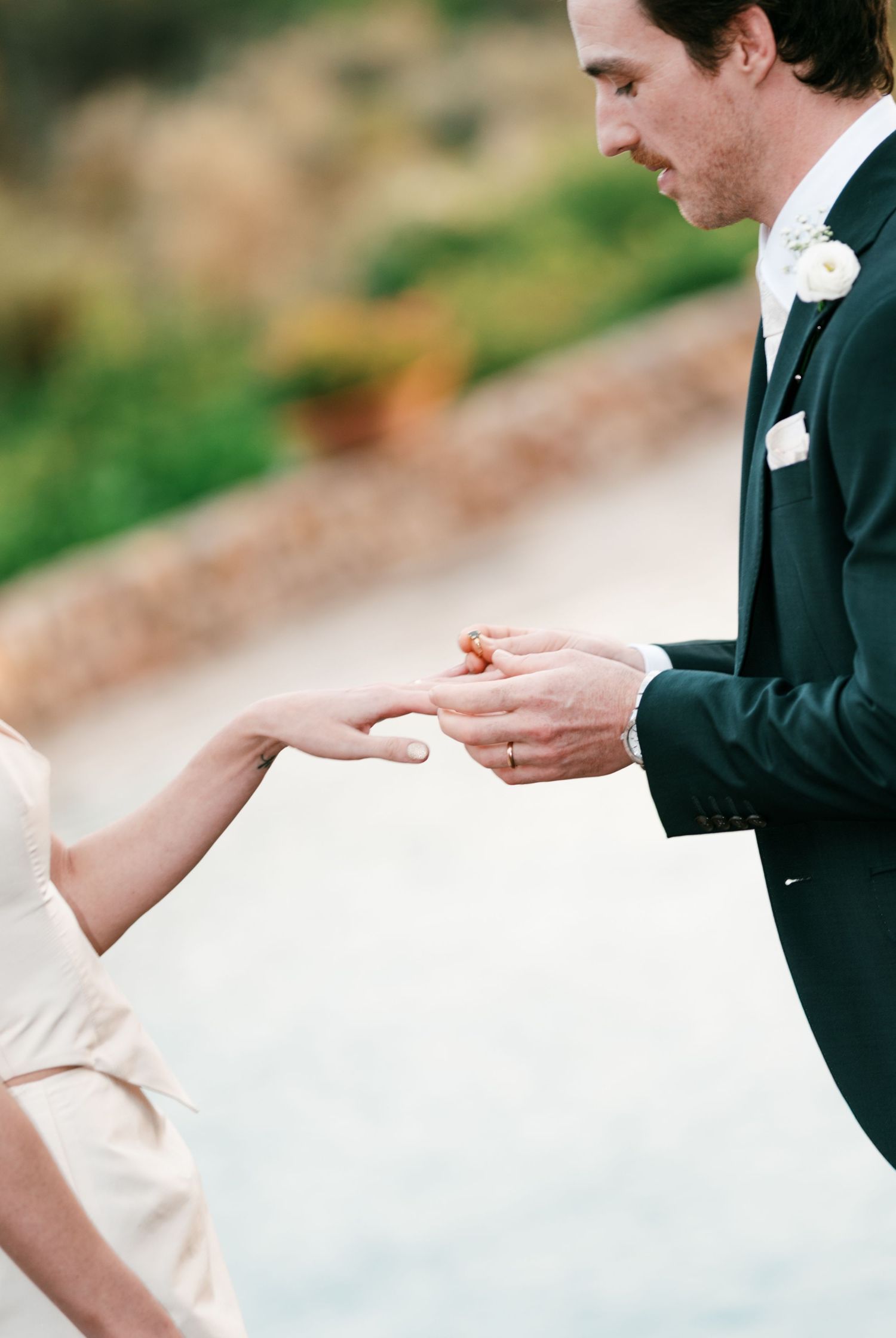 Groom in a dark suit with a white boutonniere slides a wedding ring onto his bride's finger during an outdoor ceremony.