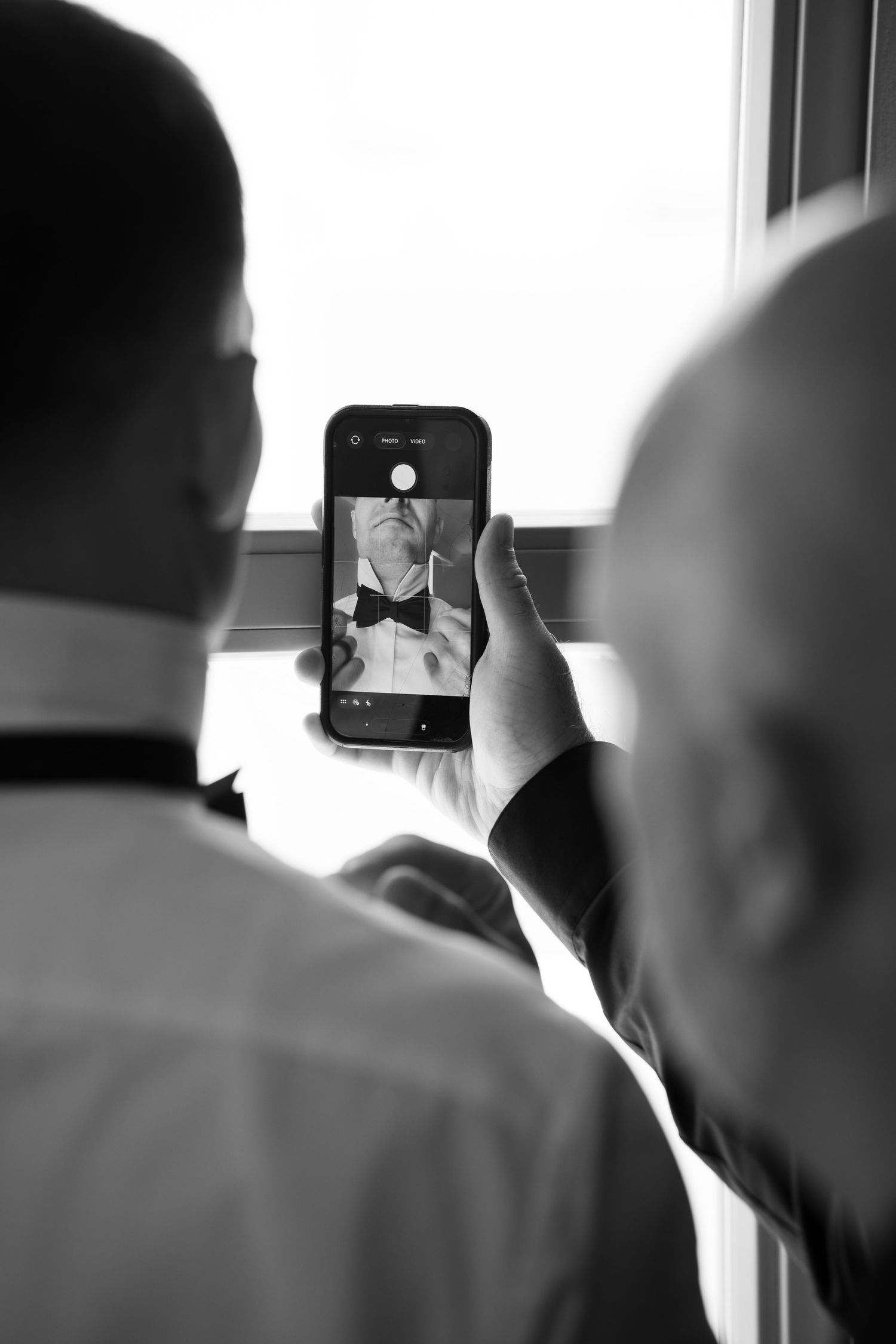 Black and white photo of two men using a smartphone to photograph a groom adjusting his bow tie before a wedding.