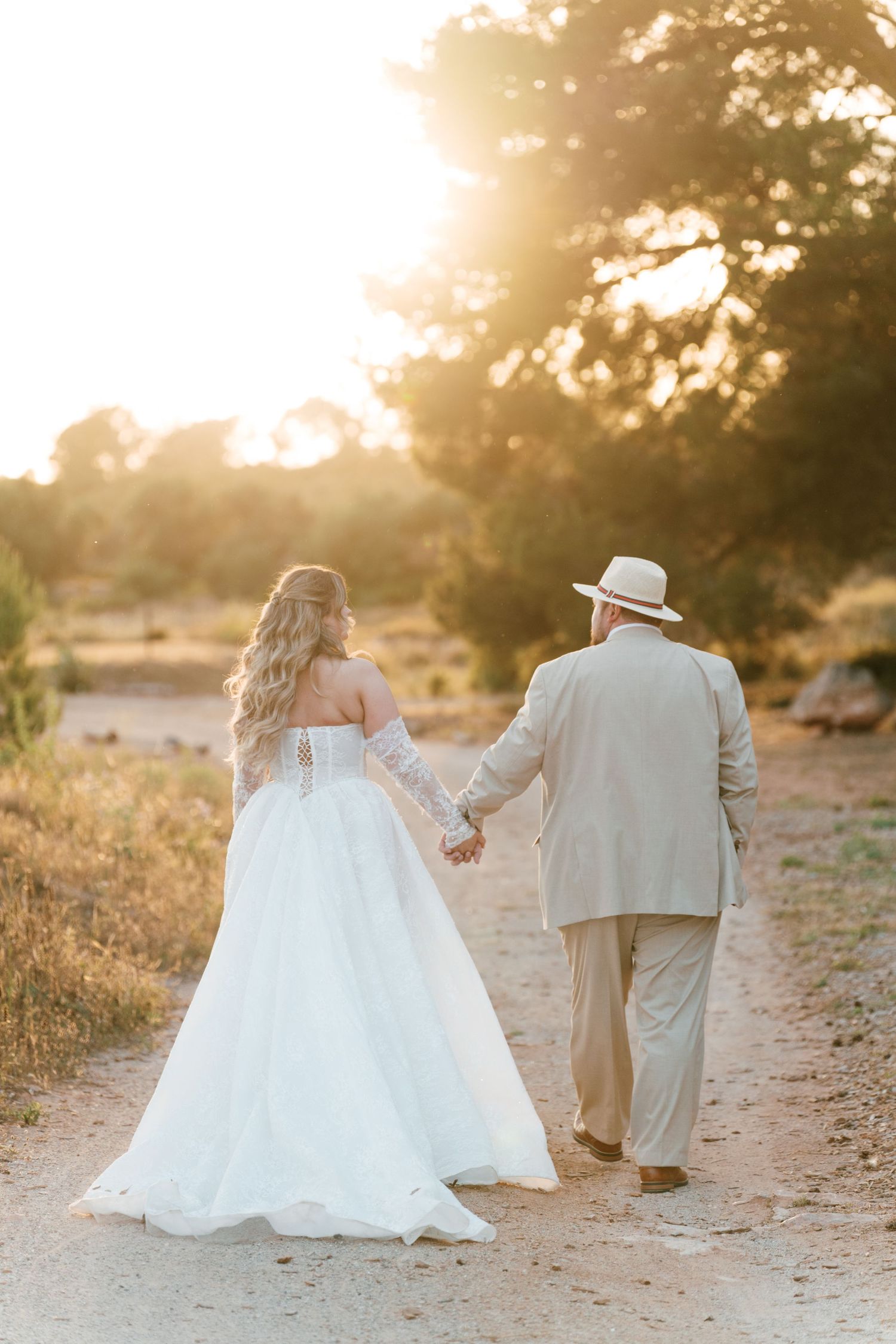 Bride and groom walk hand in hand down a golden sunlit path surrounded by trees during their wedding day.