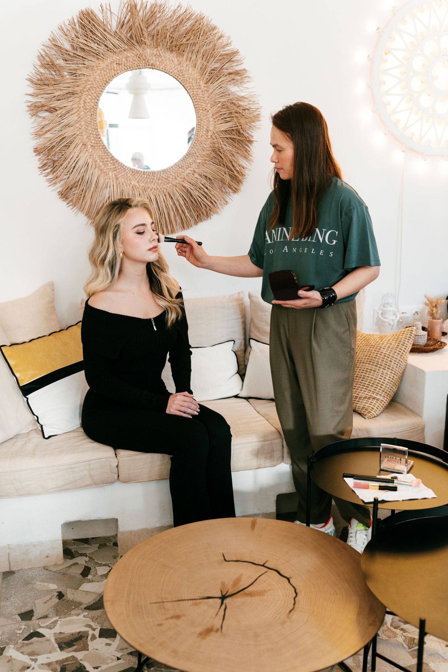 Makeup artist applying foundation to blonde woman seated on white couch in boho interior. Puglia wedding photographer, film