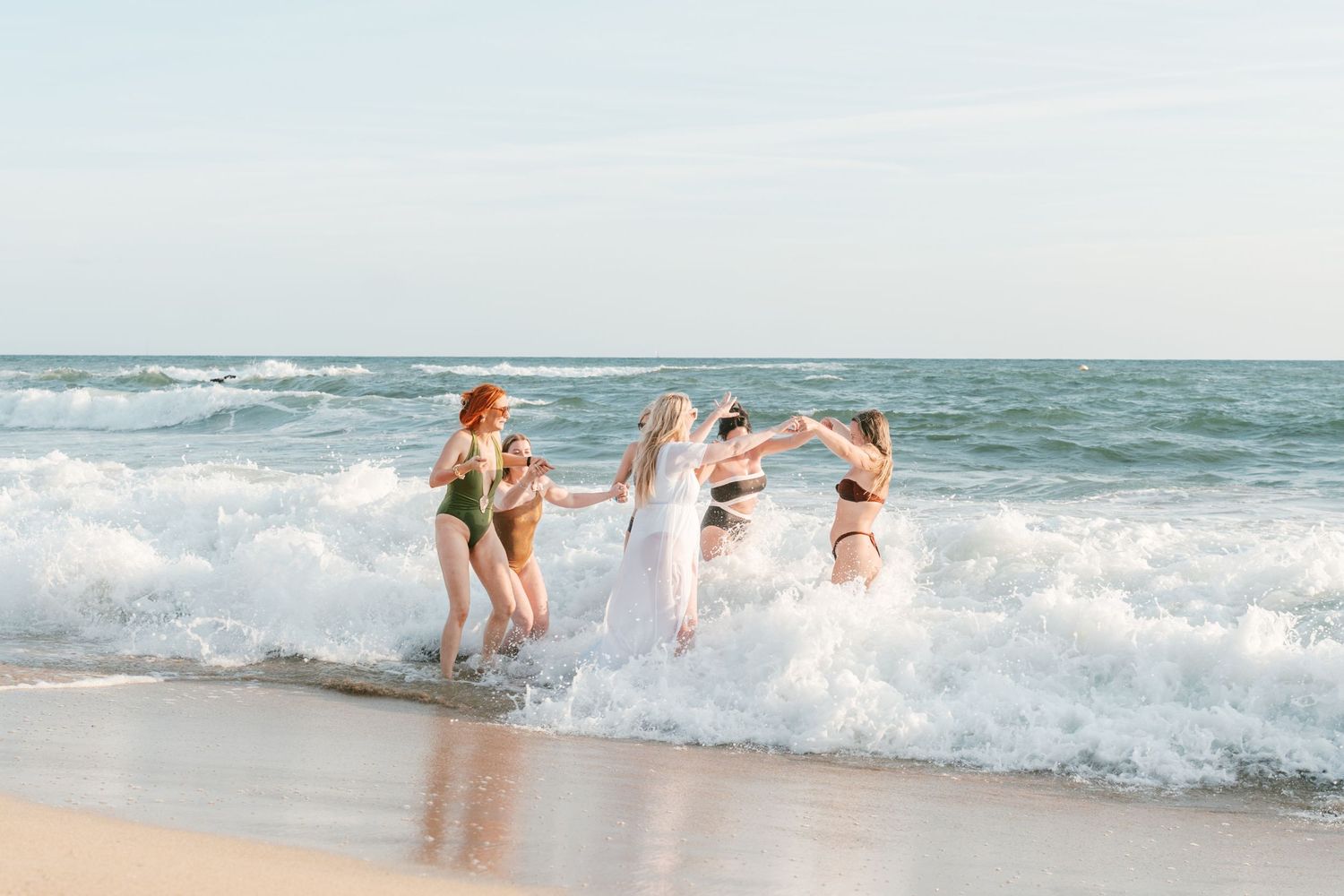 Bride in white dress celebrates with bridesmaids in swimsuits splashing in ocean waves on a sunny beach. Mallorca wedding filmmaker, video
