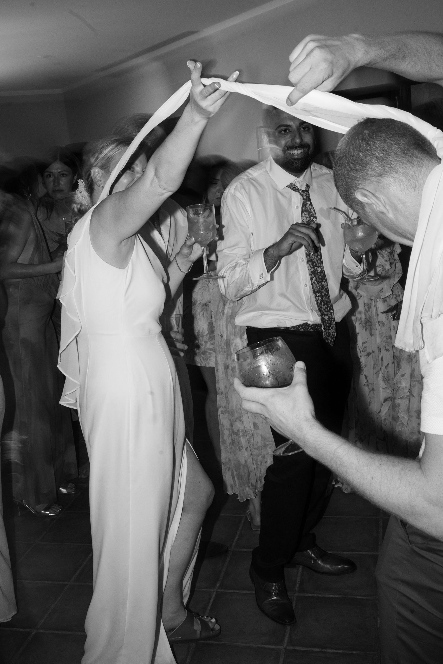 Black and white photo of wedding guests dancing under fabric arch at lively reception party. Puglia wedding photographer,