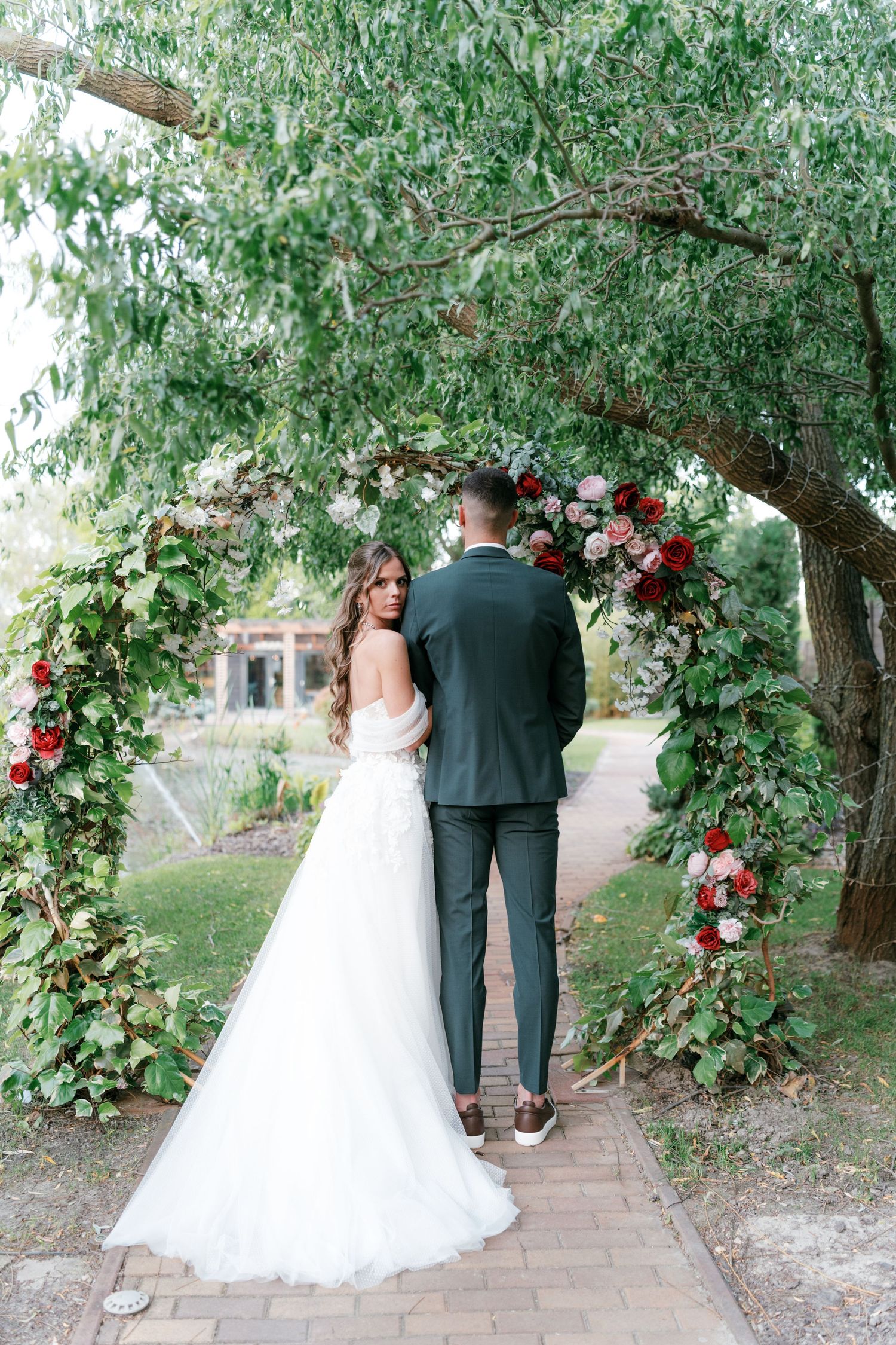 Bride and groom standing beneath floral arch adorned with red and pink roses in garden. Mallorca wedding photographer and filmmaker.